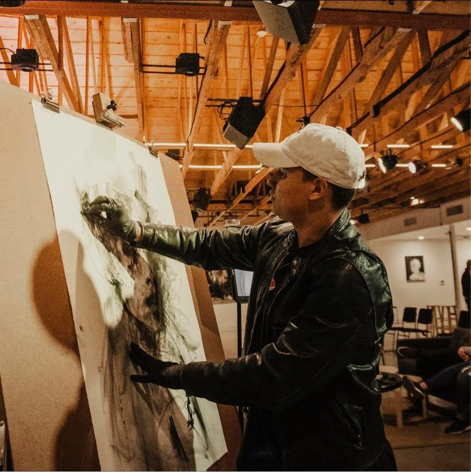 An artist wearing a white cap, black leather jacket, and gloves creating a charcoal sketch on a large easel in an art gallery with wooden ceiling beams and spotlights.
