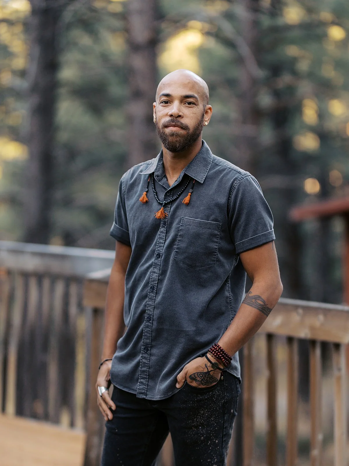 A man with a beard and shaved head wearing a dark gray short-sleeved button-up shirt, standing on a wooden outdoor deck surrounded by trees.