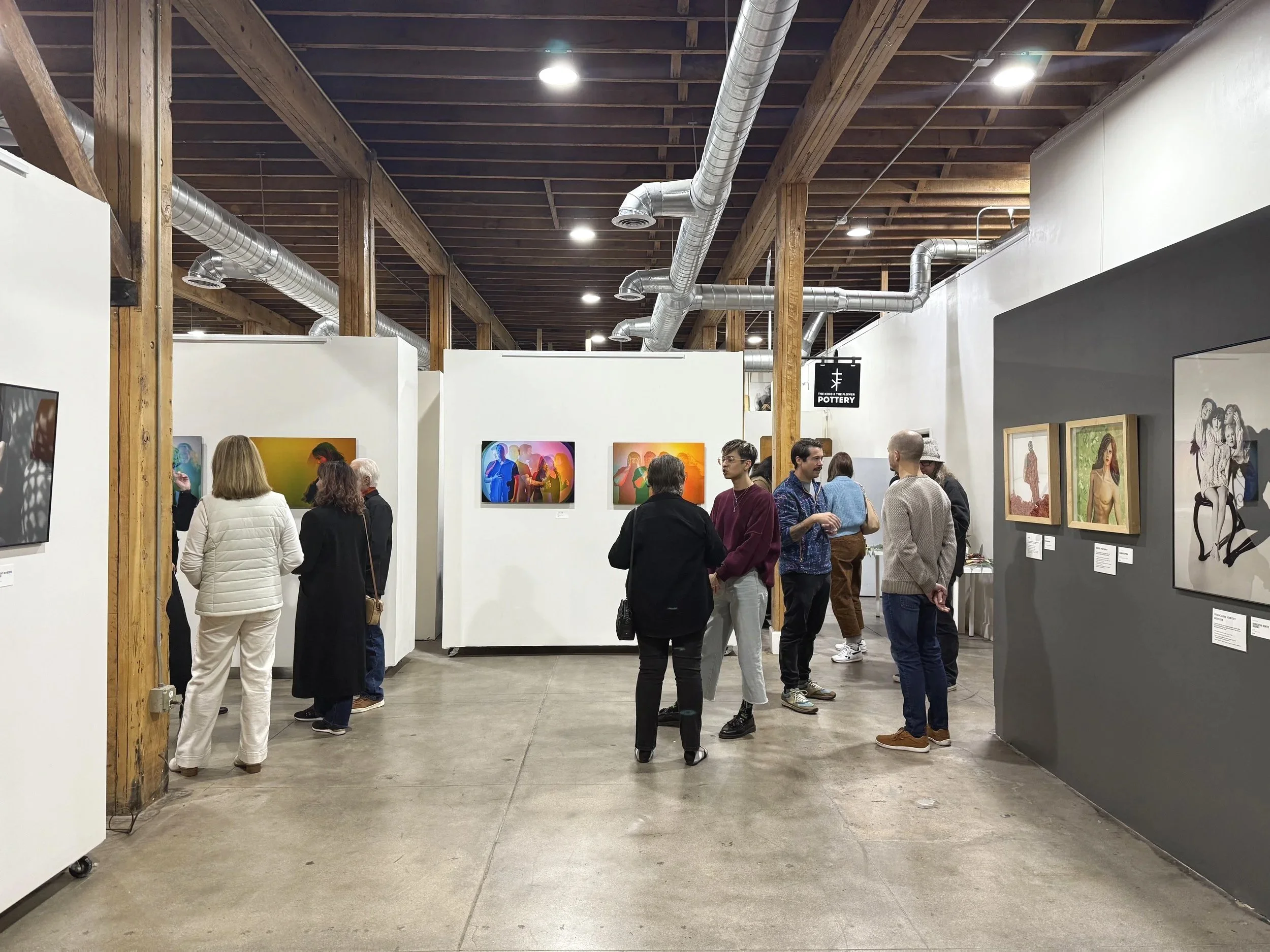 People viewing artwork in an art gallery with white and dark gray walls, wooden beams, and exposed ductwork on the ceiling.