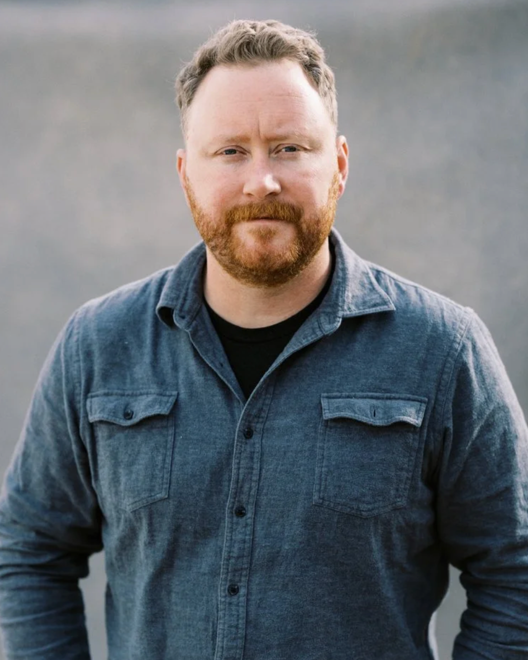 A man with a red beard, short curly hair, wearing a denim jacket, standing outdoors with blurred background.