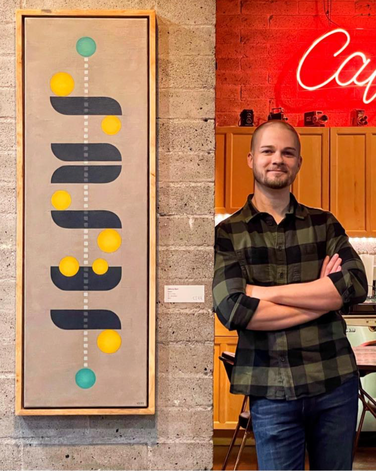 A man with a beard and short hair standing with crossed arms in front of a brick wall, next to a framed abstract artwork featuring a vertical line with yellow and teal circles and black curved shapes, in a cafe setting with wooden cabinets and neon sign.
