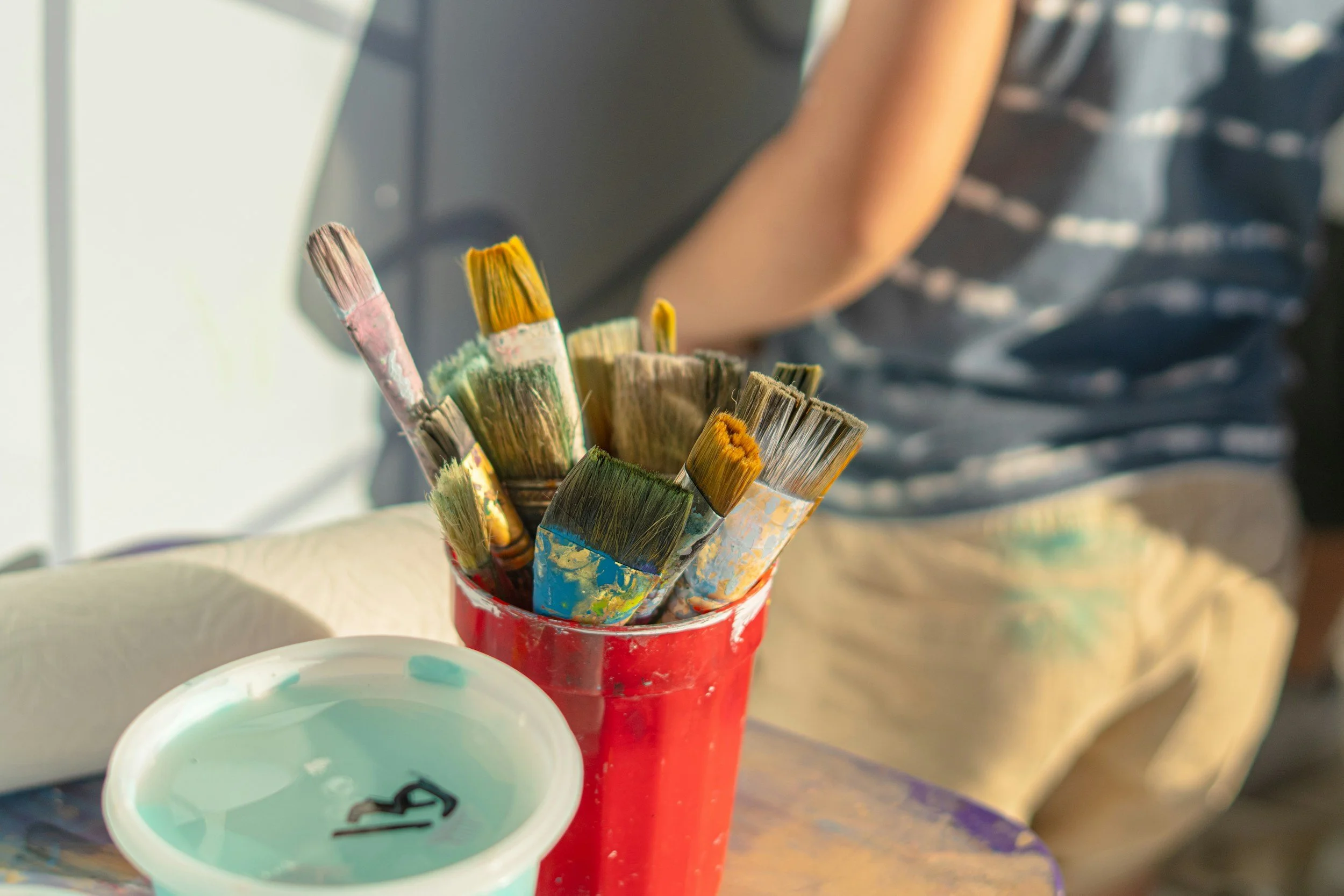 Collection of used paintbrushes in a red container with a small bowl of blue-green paint nearby.