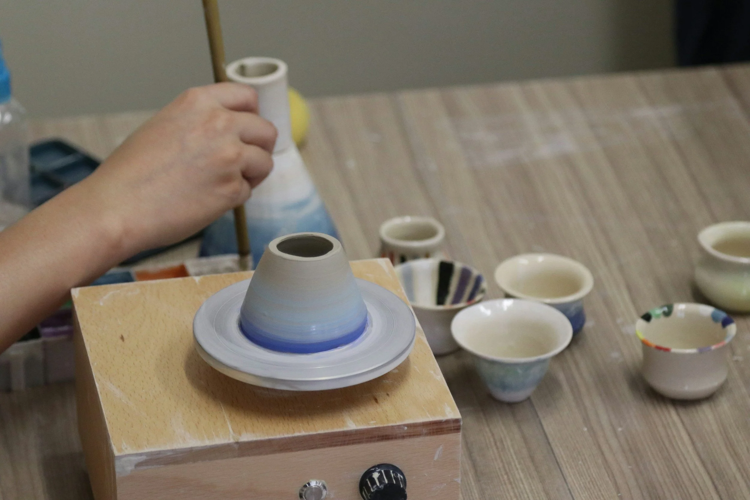 Hands shaping a ceramic bowl on a pottery wheel surrounded by several finished painted bowls on a wooden table.