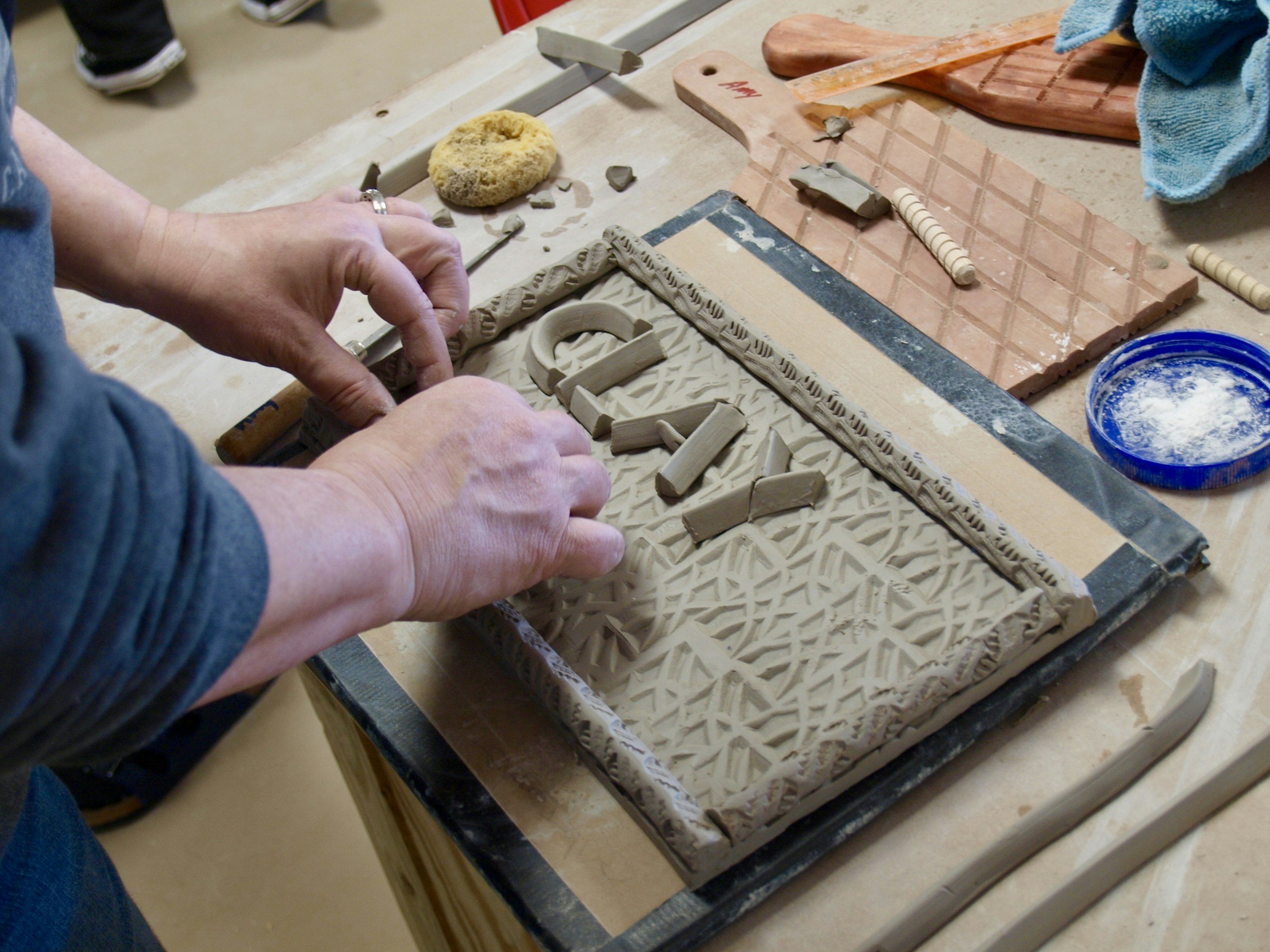 Person working with clay on a slab at a pottery or sculpture studio, surrounded by tools and clay pieces.