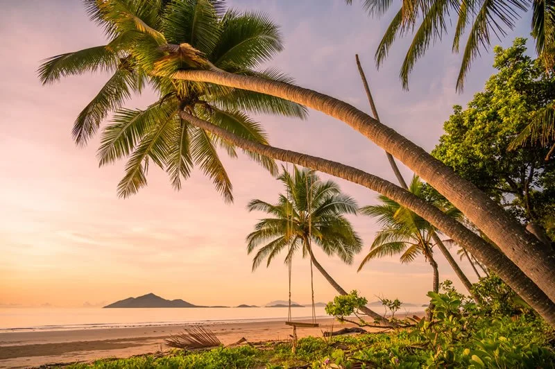 Sunset view on a tropical beach with palm trees leaning over the sand, and small islands in the distance.