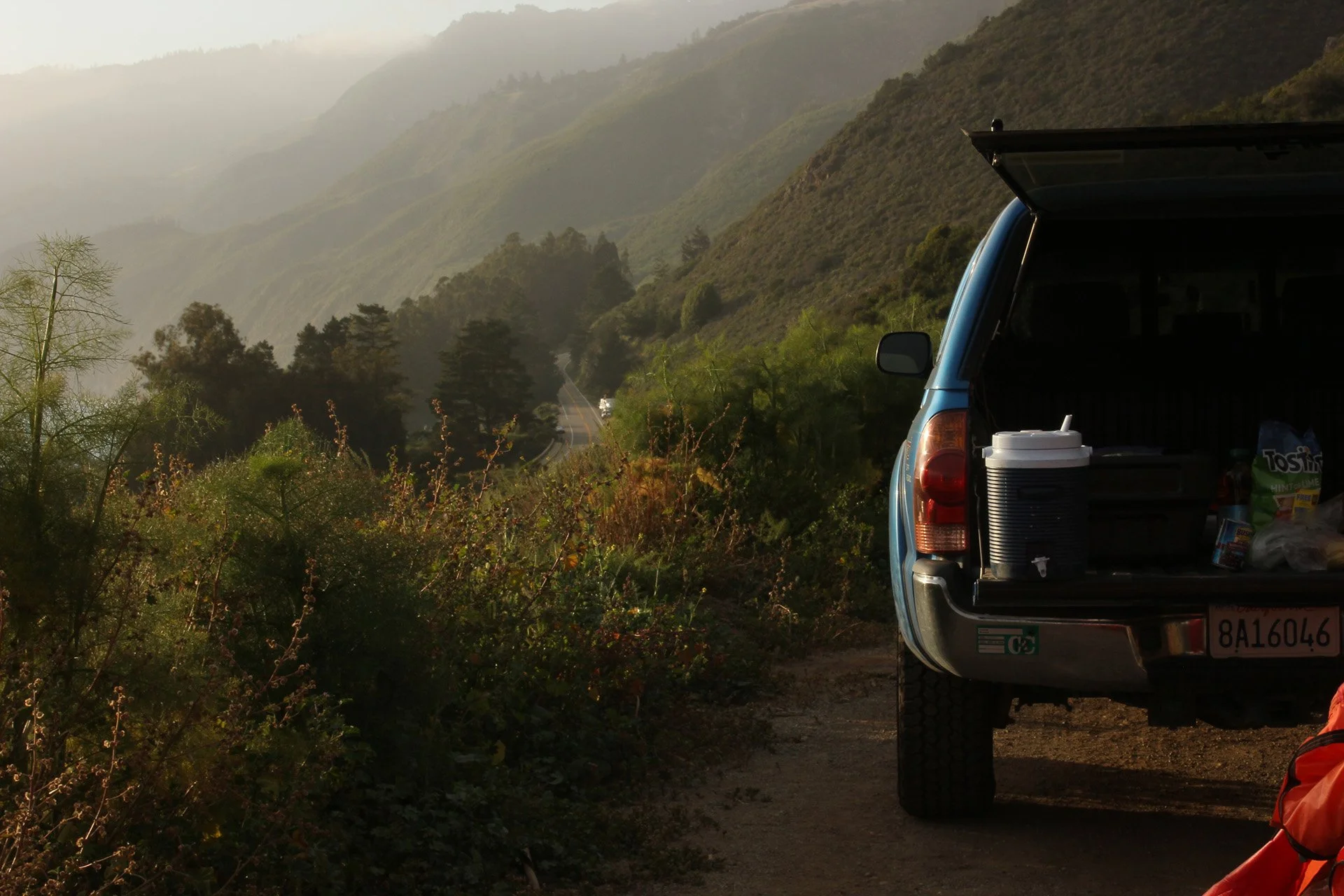 A blue truck is parked by the side of the highway, the tailgate down with camping supplies in the back. The highway leads north into the mountains. The Pacific Ocean is to the left of the highway.