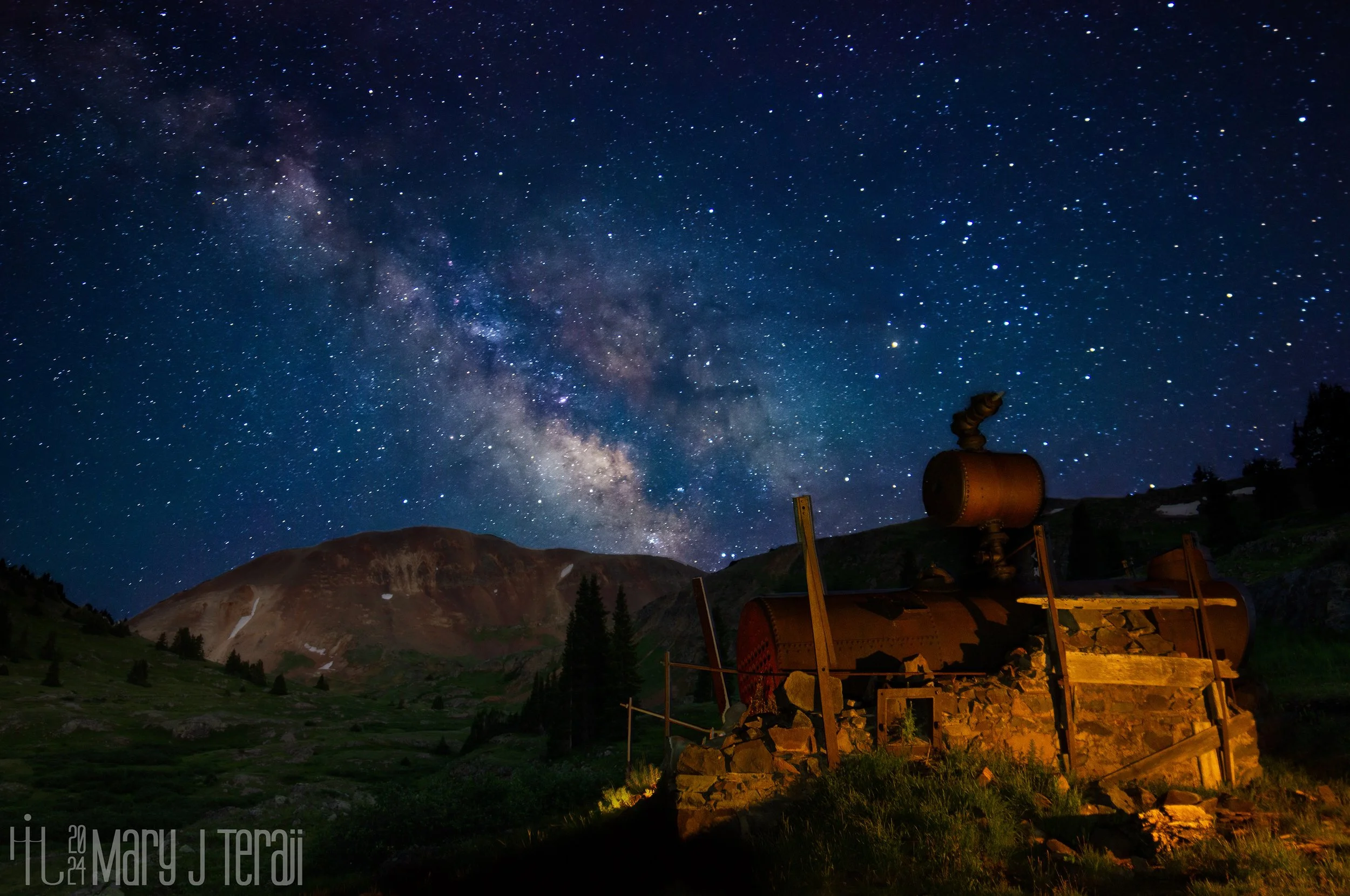 The Milky Way stretches across a starry night sky above an old rusted boiler and stone structure, set against a rugged mountain landscape.