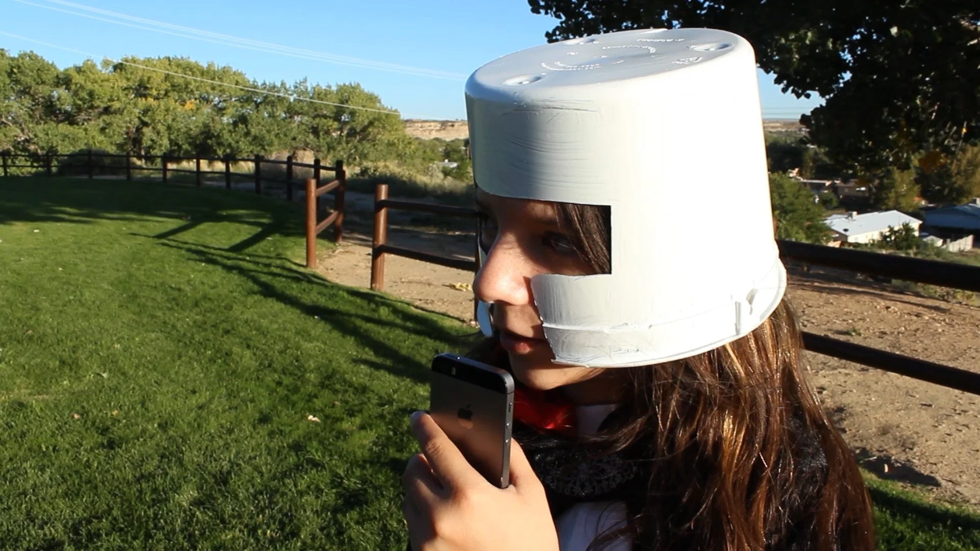 A little girl stands in a grassy field wearing a bucket as a space helmet.
