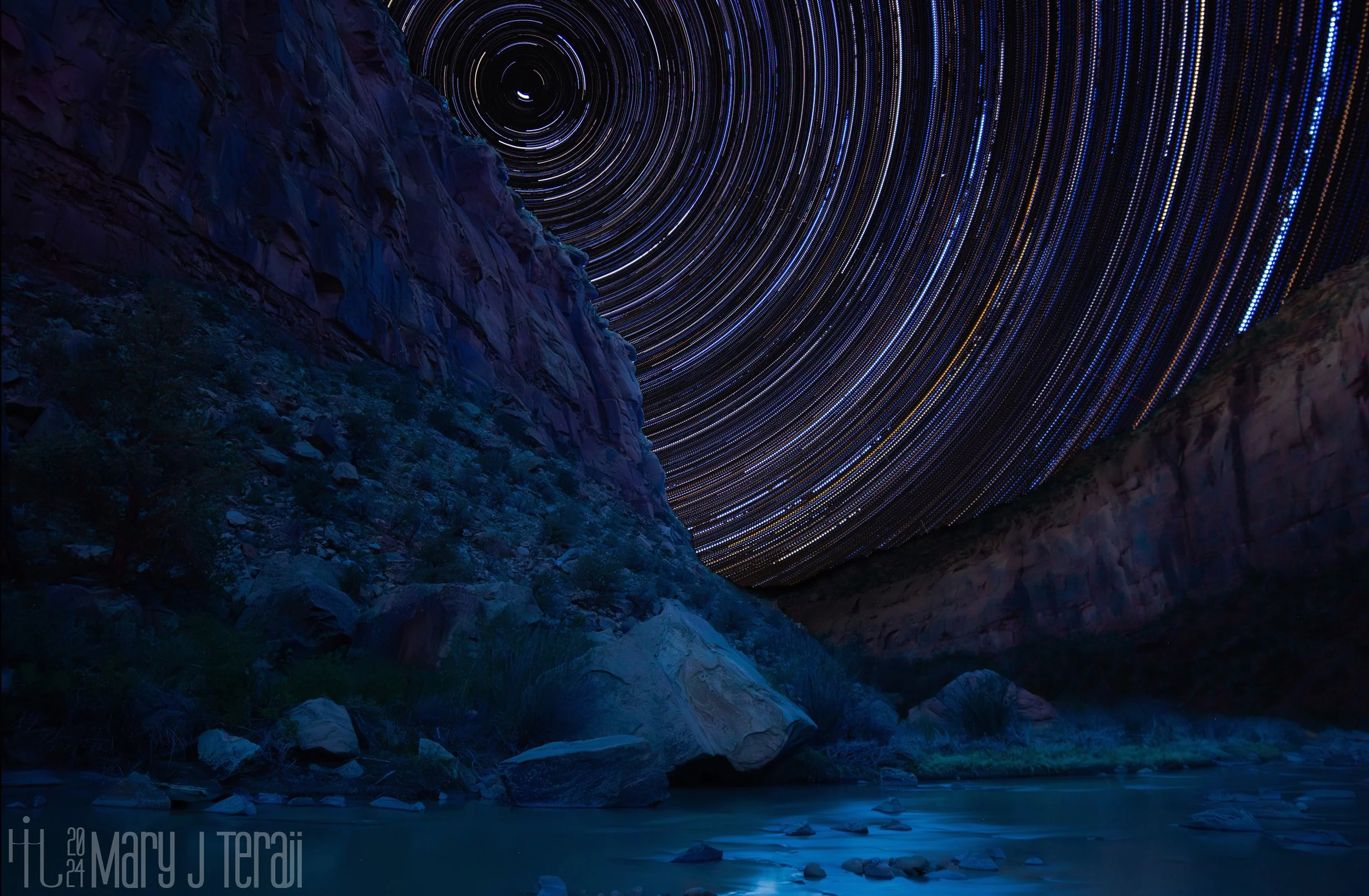 Long-exposure star trails swirl above a rugged canyon at night, creating a hypnotic vortex of light against towering rock formations and a river below.