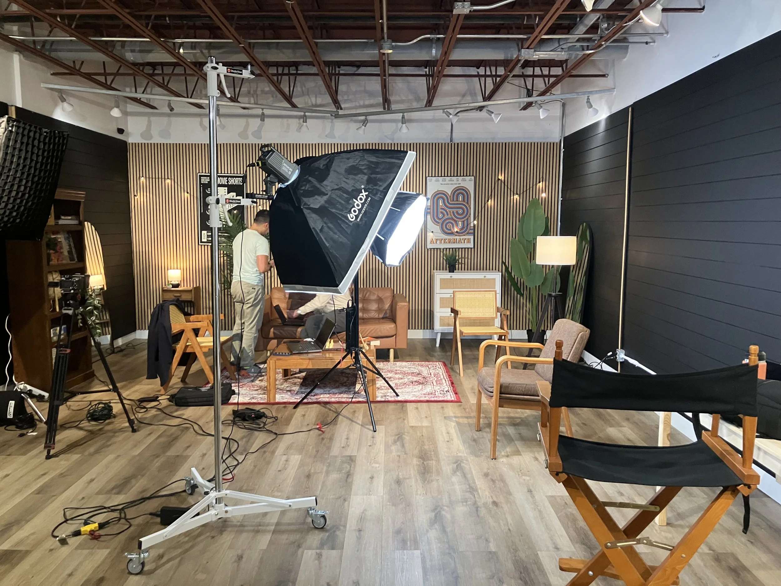 A photography studio with lighting equipment, a camera on a tripod, and a person working on a laptop. The studio has a sofa, chairs, a rug, and decorative plants, with a wooden slat wall and posters on the wall.