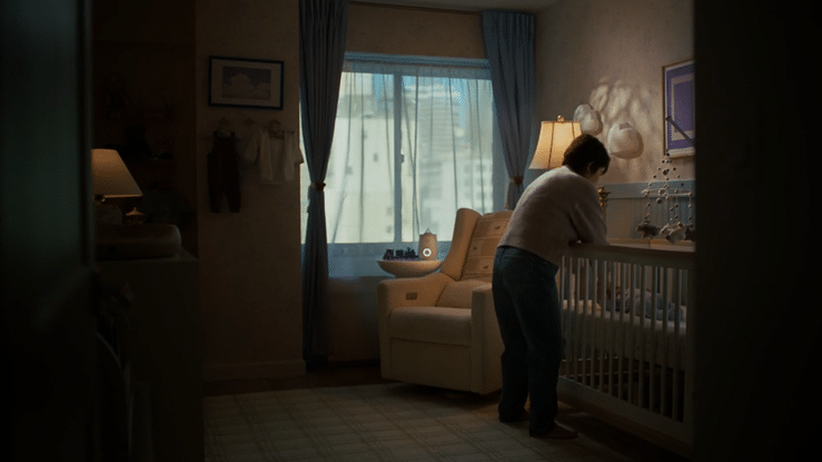 A woman leaning over a crib inside a cozy bedroom with blue curtains, a beige armchair, and warm lighting.
