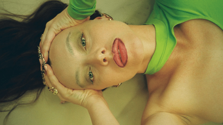 A woman lying on her back with her head tilted, wearing a bright green top, with her hands holding her head, and making a playful facial expression with her tongue out and eyes half-closed.