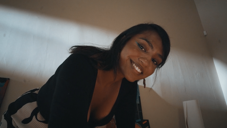 A woman smiling and leaning forward, with long dark hair, wearing a black top and striped pants, sitting indoors near a wall and a lamp.