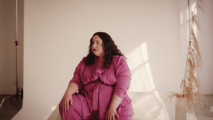 A woman with dark curly hair wearing a pink dress sitting on a white surface in a room with a plain beige wall and dried plants on the right side.