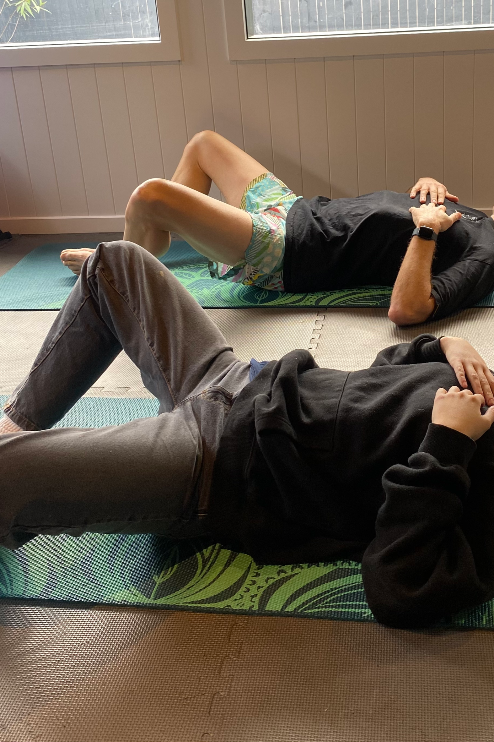 Two people lying on exercise mats in a room with large windows, one wearing a black hoodie and jeans, the other in a black t-shirt and shorts, relaxing or stretching.