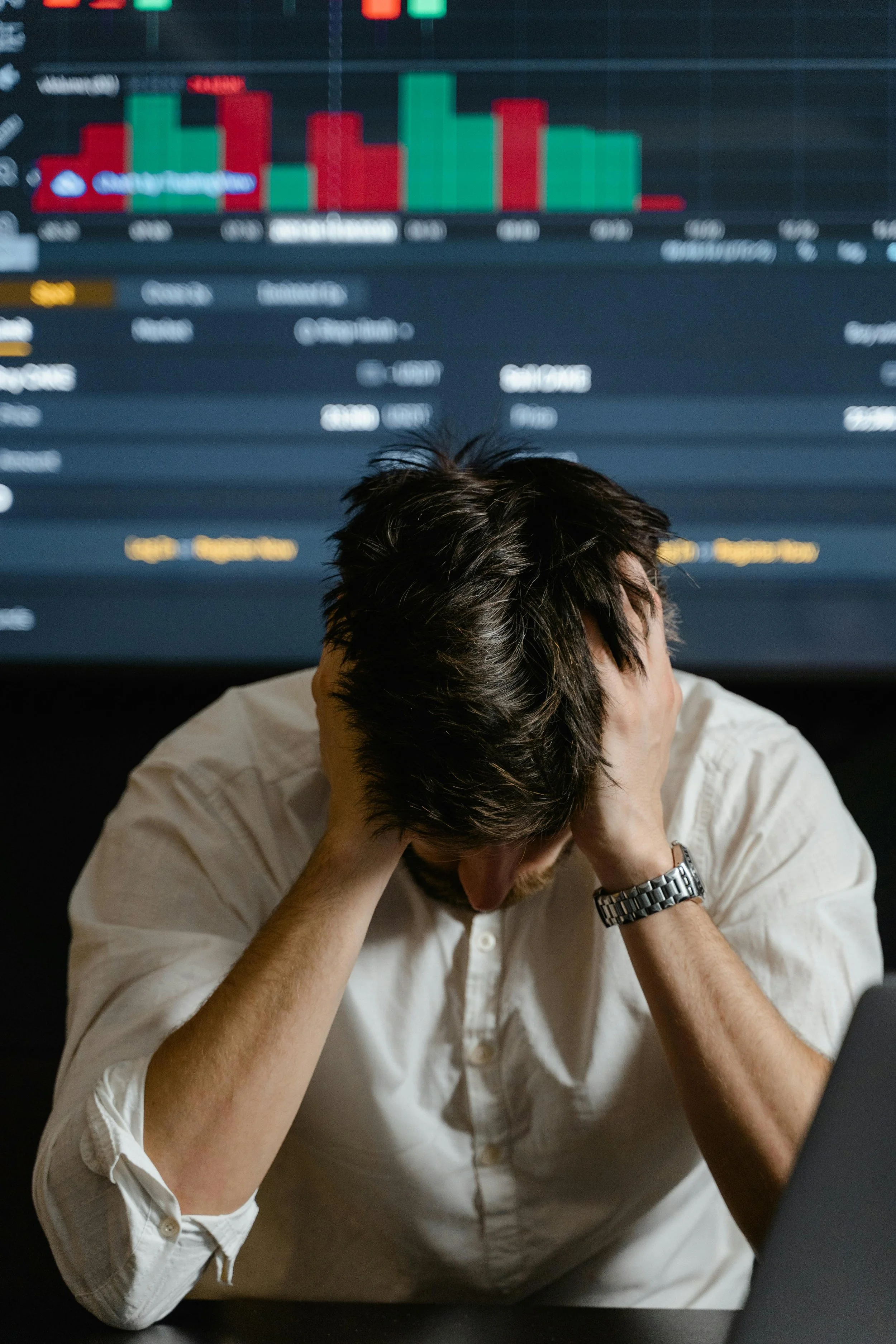 Frustrated man with hands on his head in front of a large screen displaying stock market graphs and financial data.