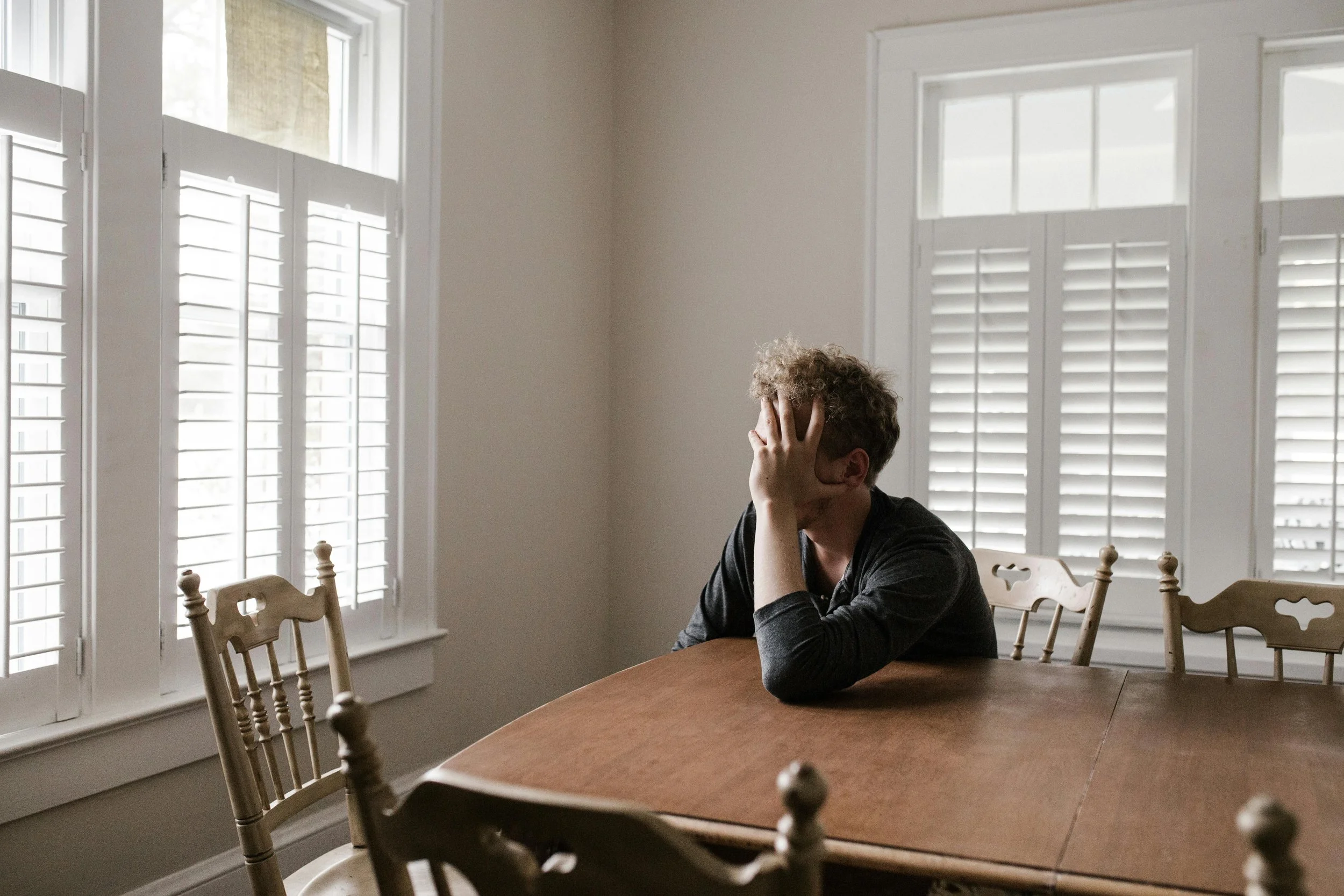 A man sitting at a dining table with his face in his hand, appearing distressed or upset, in a room with white walls and large windows with white shutters.