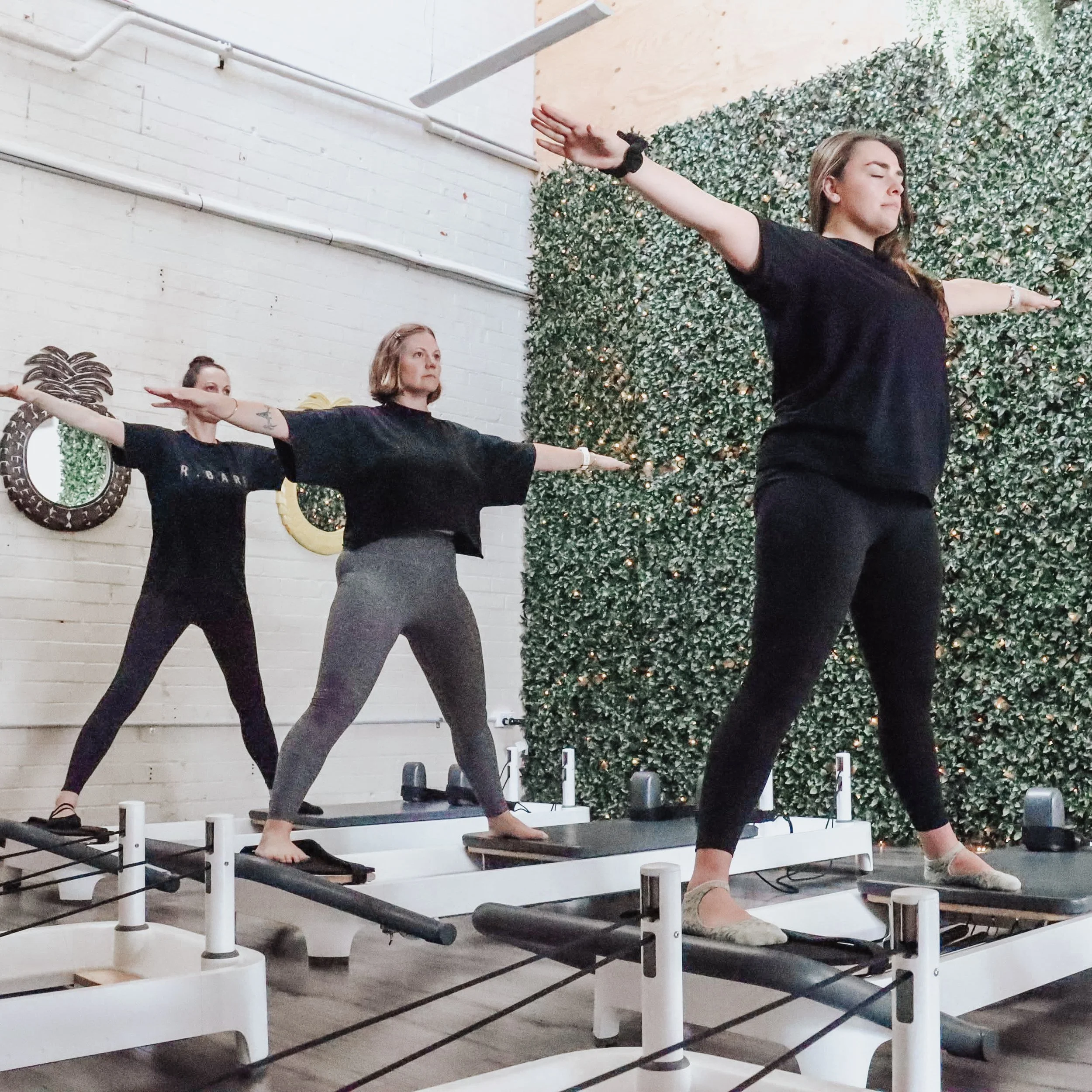 Three women participating in a reformer Pilates class, standing on reformer machines with their arms outstretched in a T-shape, in a studio with a white brick wall and green foliage wall.