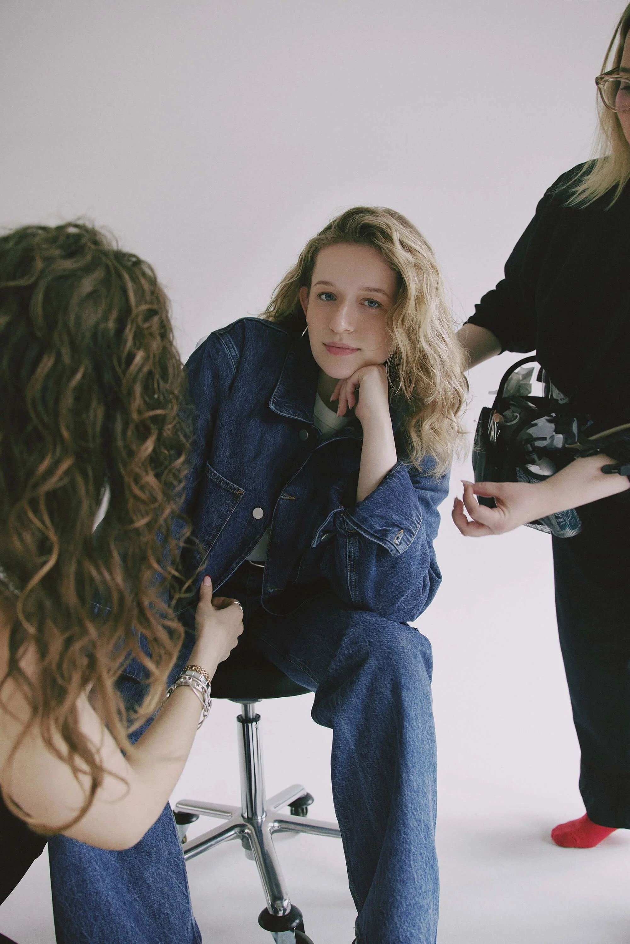 Junge Frau sitzt auf einem Stuhl, während zwei Frauen auf sie zukommen, eine mit Locken und die andere mit blonden Locken, im Fotostudio.