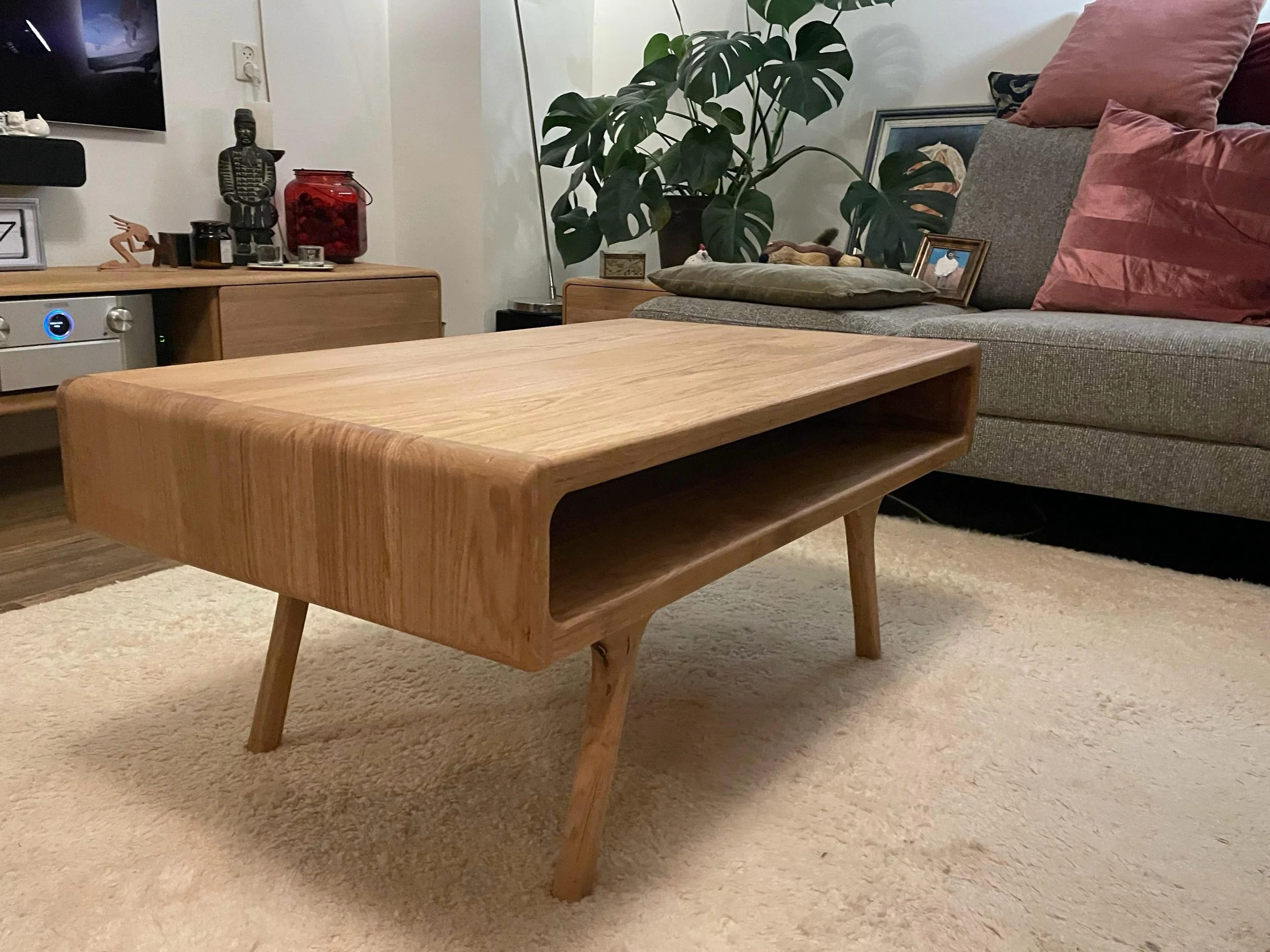 Een eikenhouten design salontafel op een beige tapijtvloer in een woonkamer, met een bank met roze en grijze kussens, een plant, en decoratieve objecten op een kast in hetzelfde design op de achtergrond.