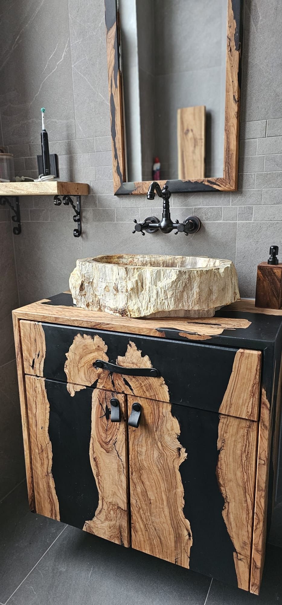 Een badkamermeubel met een houten kast en een natuurstenen wastafel, een spiegel met een houten frame en een zwarte kraan. Op de kast staat een zeepdispenser.