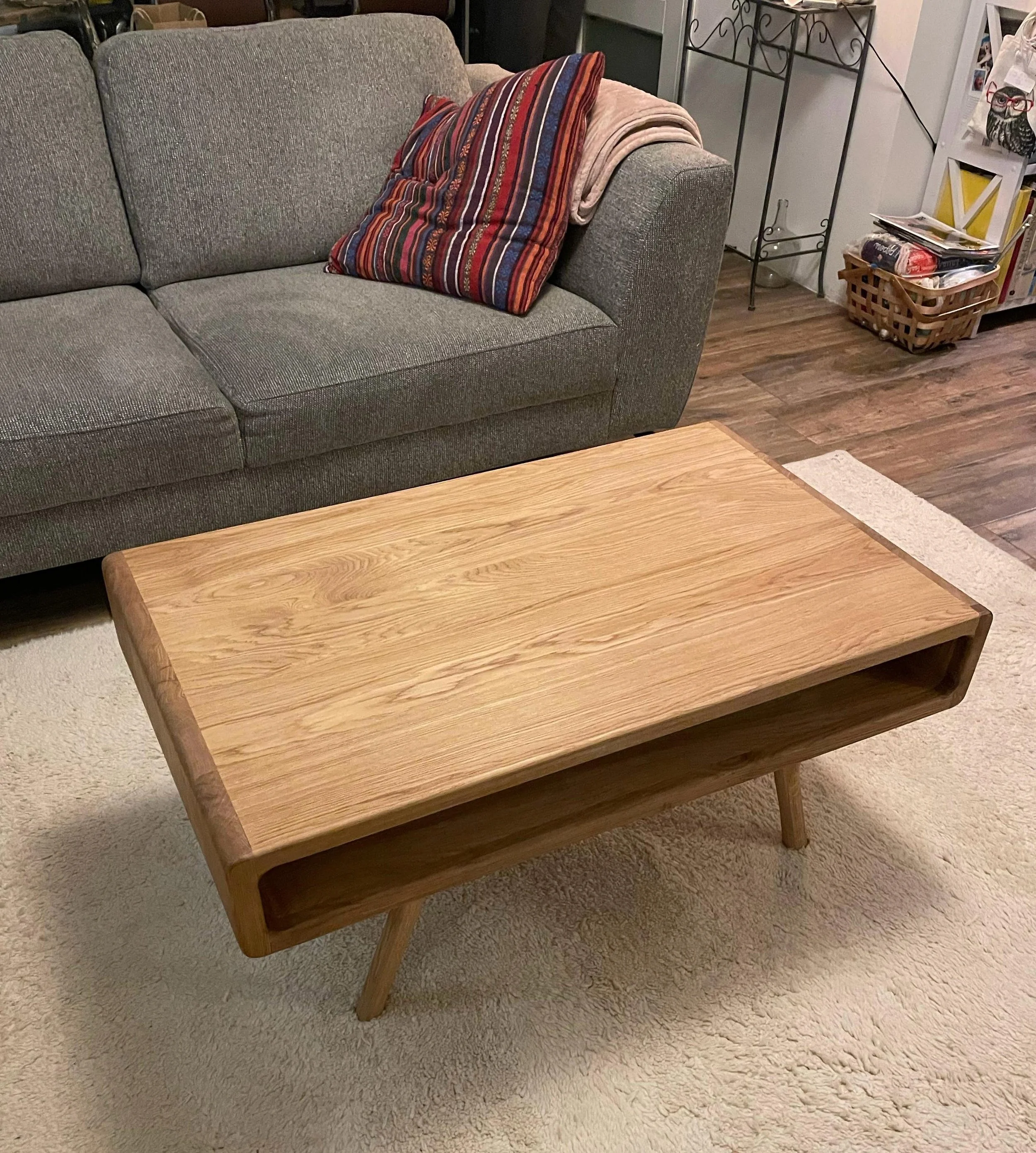 Een houten moderne salontafel op een beige tapijt in een woonkamer, met een grijze bank en kussens op de achtergrond.