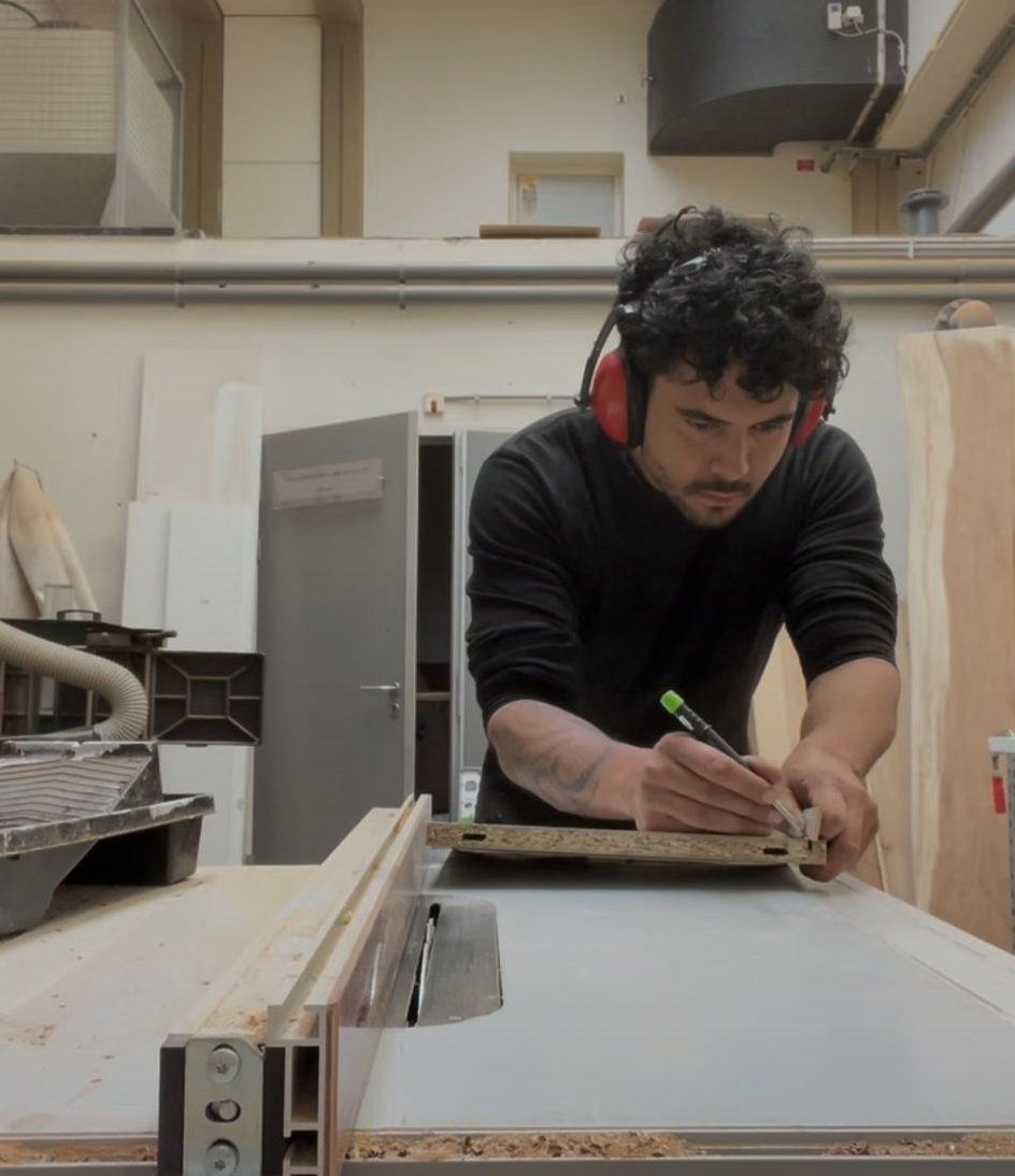 Man die in een houtwerkplaats werkt, met geluiddempende koptelefoon op, die een stuk hout of MDF onder een randlijn op een werktafel markeert met een potlood.