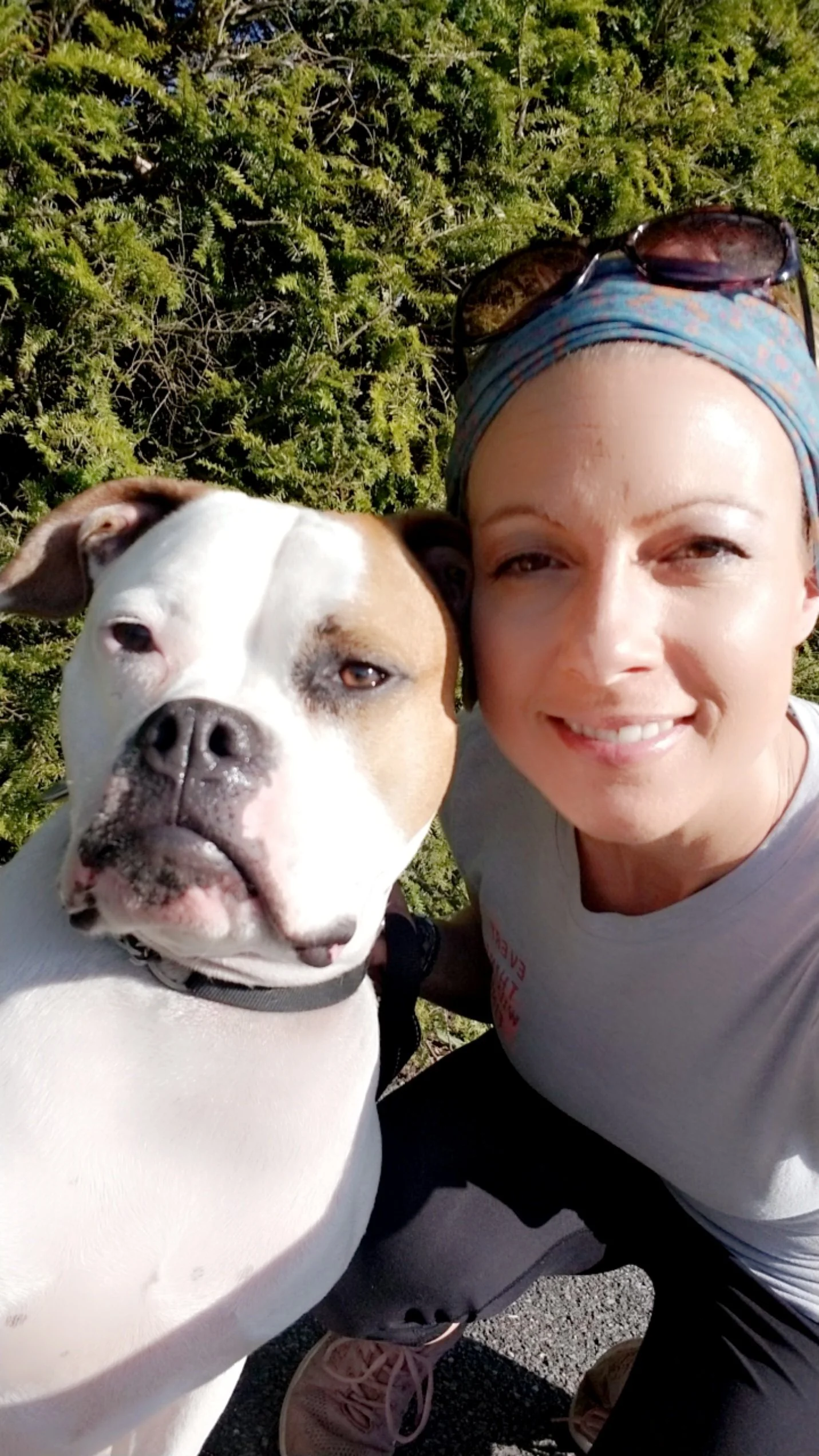 A woman taking a selfie with a large white and brown dog outdoors, with greenery in the background. The woman is smiling, wearing a blue headscarf with sunglasses on top, and white shirt. The dog has a black collar and is looking at the camera.