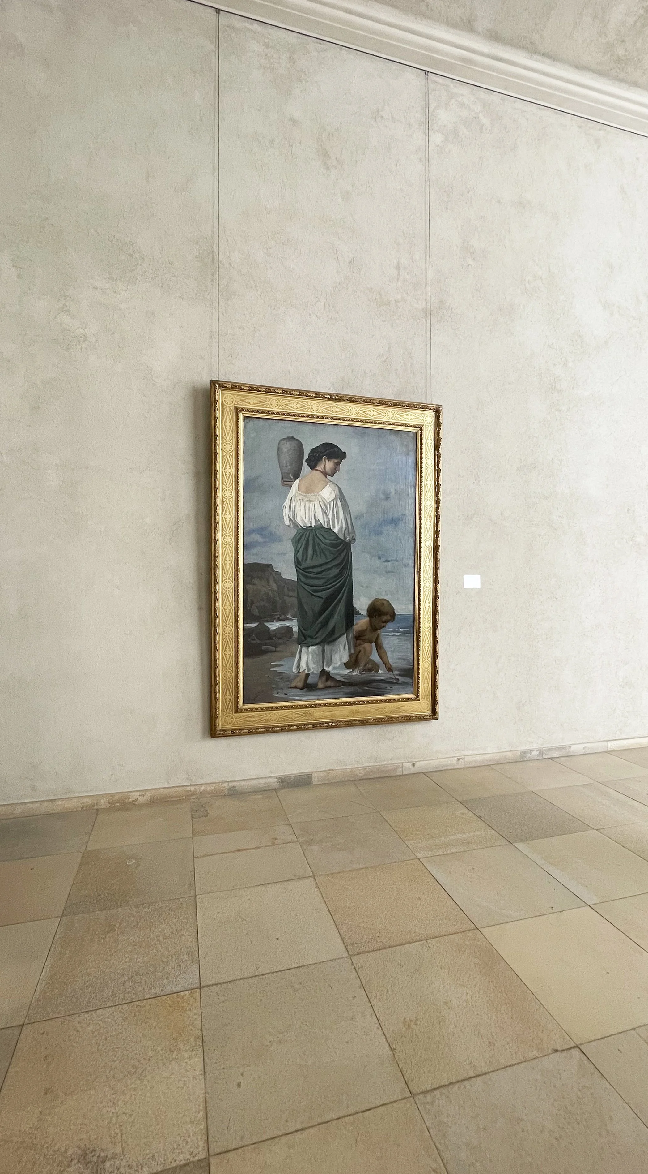 Painting of a woman with dark hair in a white and green dress standing on the beach with a small child, with a rocky shoreline and cloudy sky in the background, framed in an ornate gold frame.