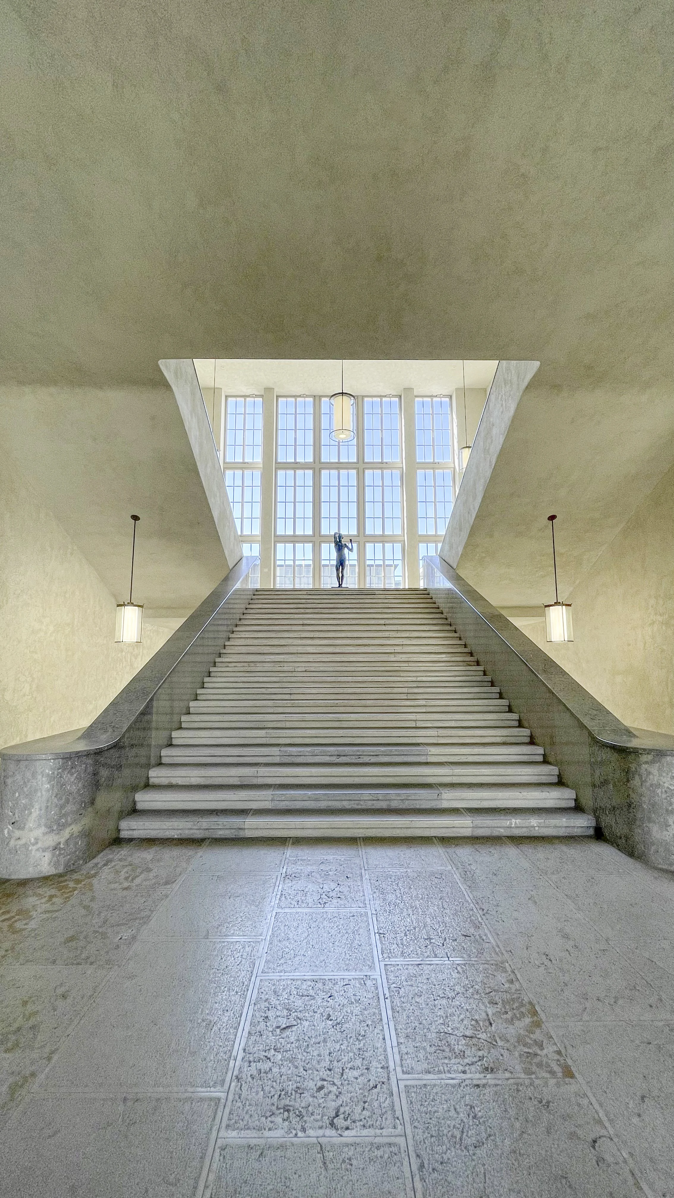 Interior view of a grand staircase leading up to a large window with a sculpture of a person holding an object at the top, flanked by two hanging lamps.