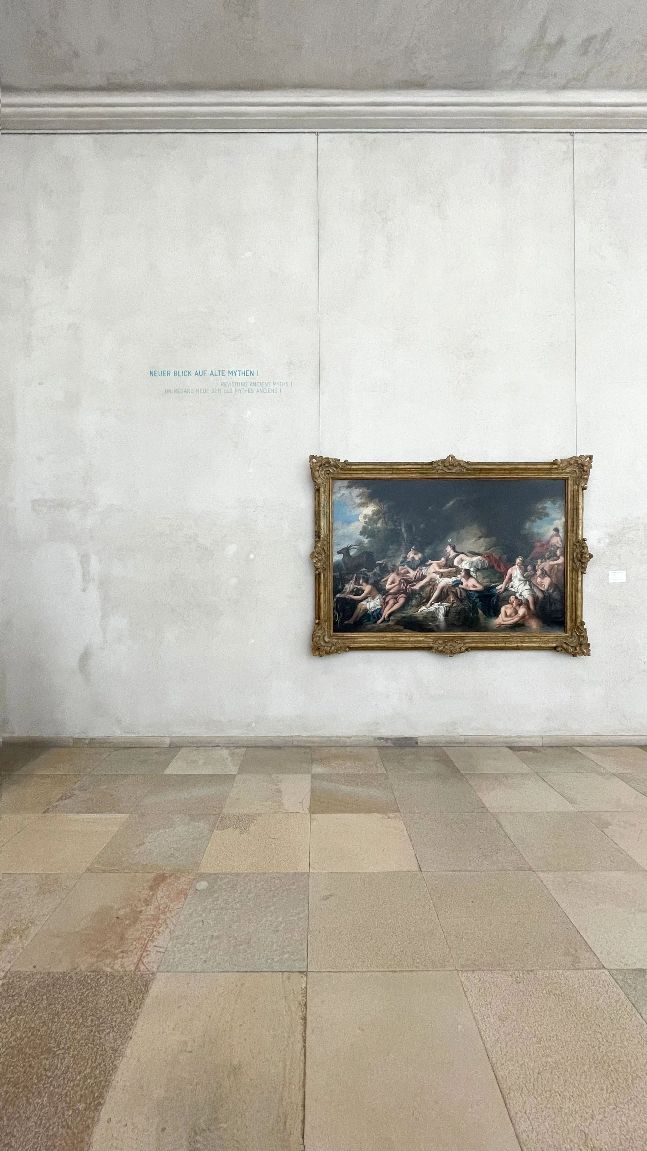 A painting in an ornate frame on a gallery wall, with a light-colored stone floor and a plain wall displaying text in German, French, and English.