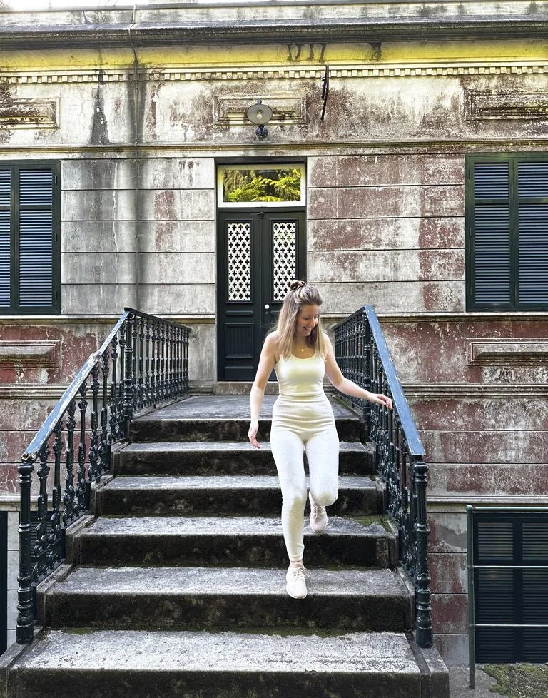 Woman in white outfit descending outdoor stone staircase with black metal railings, in front of a weathered building with green shuttered windows and a black door.