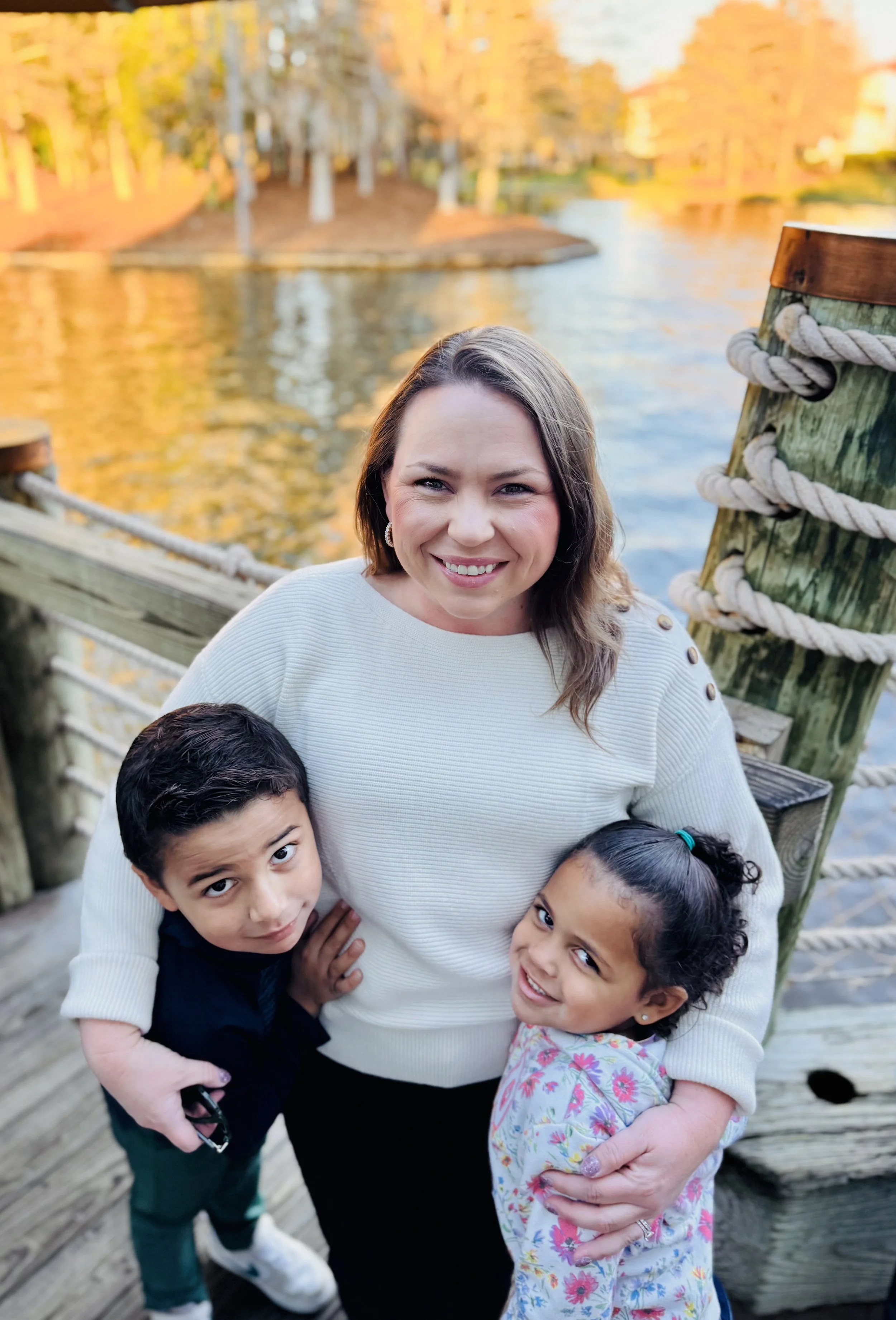 A woman with two children smiling at the camera on a wooden dock by a river with fall foliage.