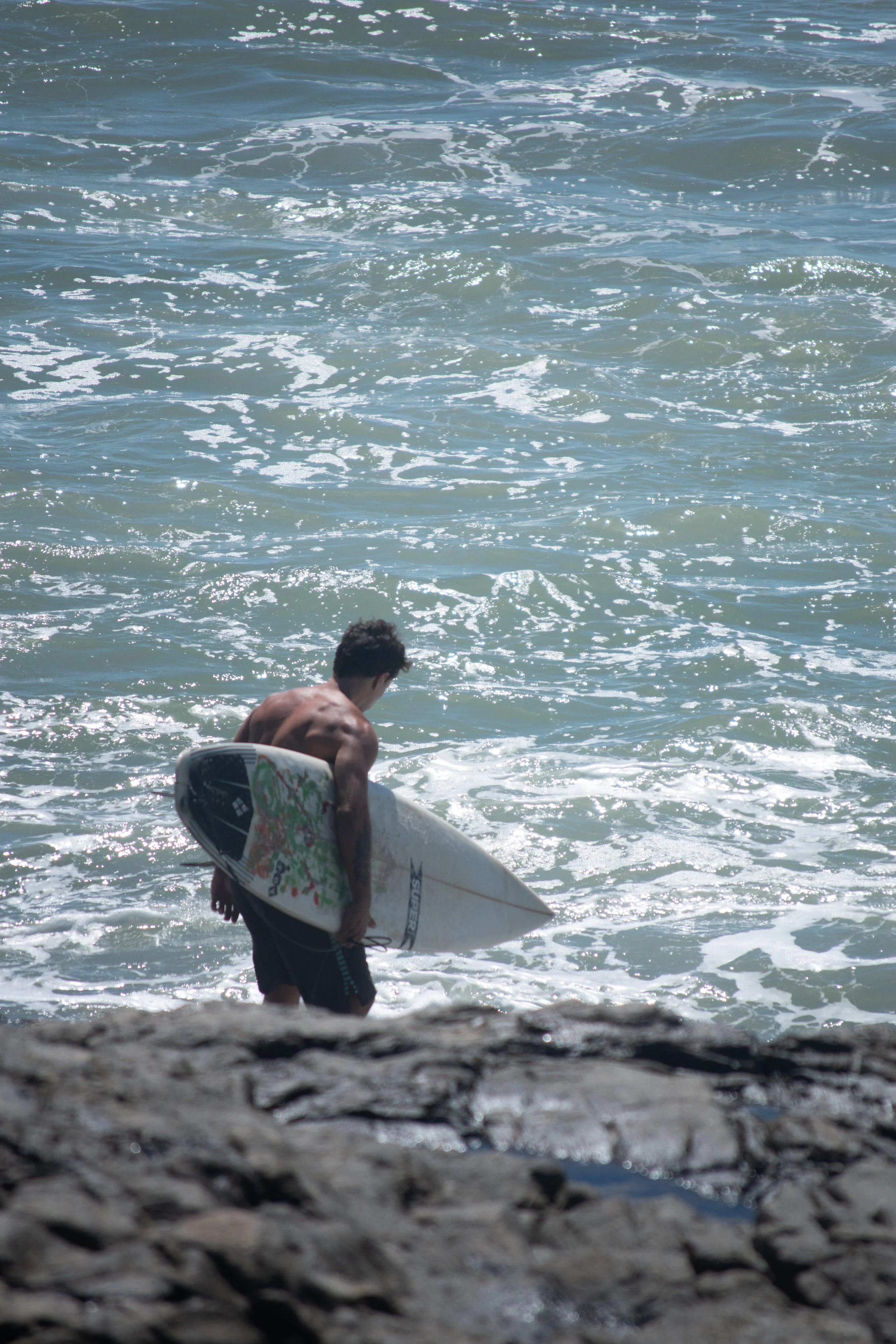 A man carrying a surfboard walking into the ocean from a rocky shoreline.