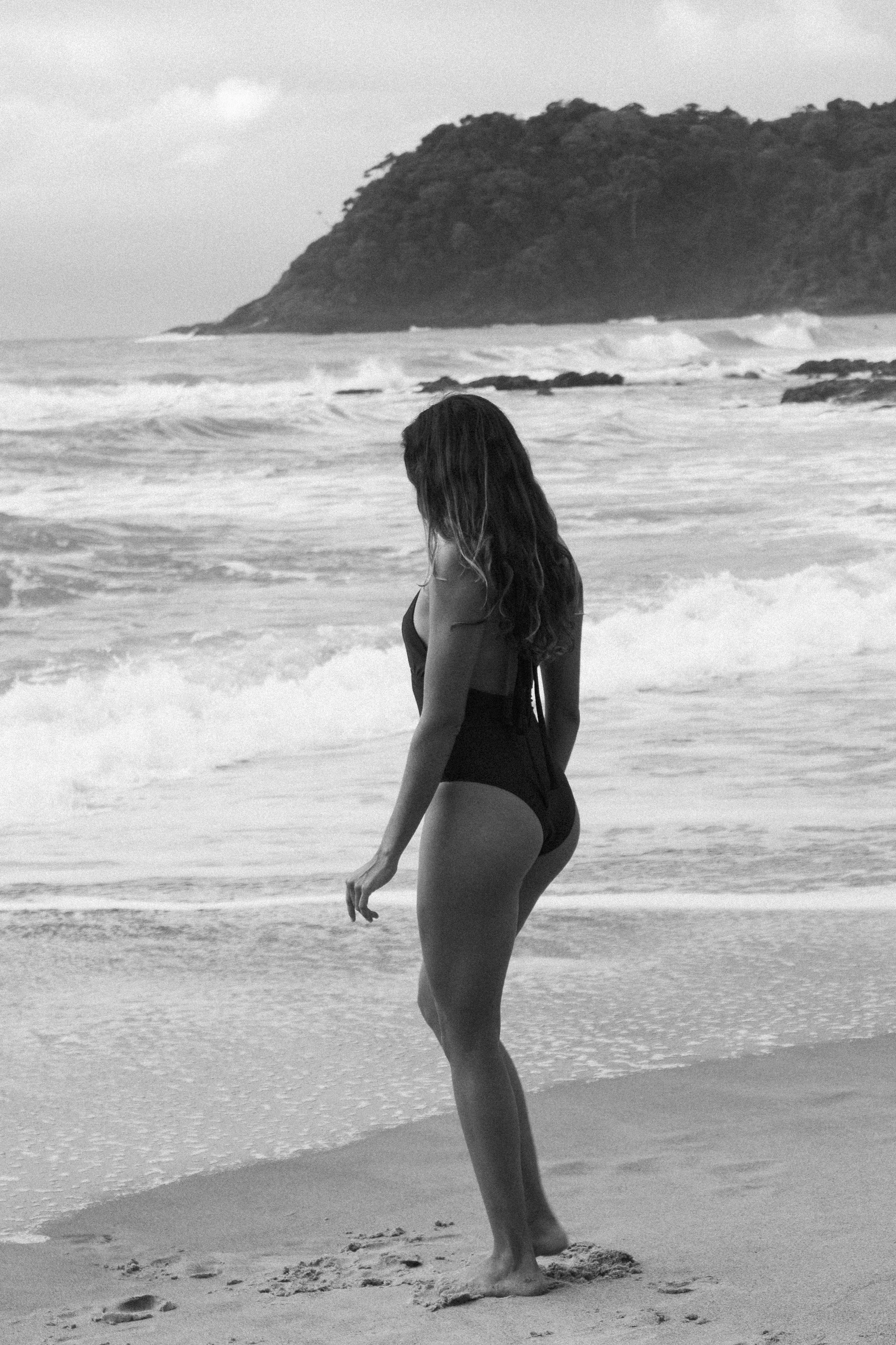 A woman standing on the beach near the shoreline, facing away from the camera, with an island or hillside in the distance.