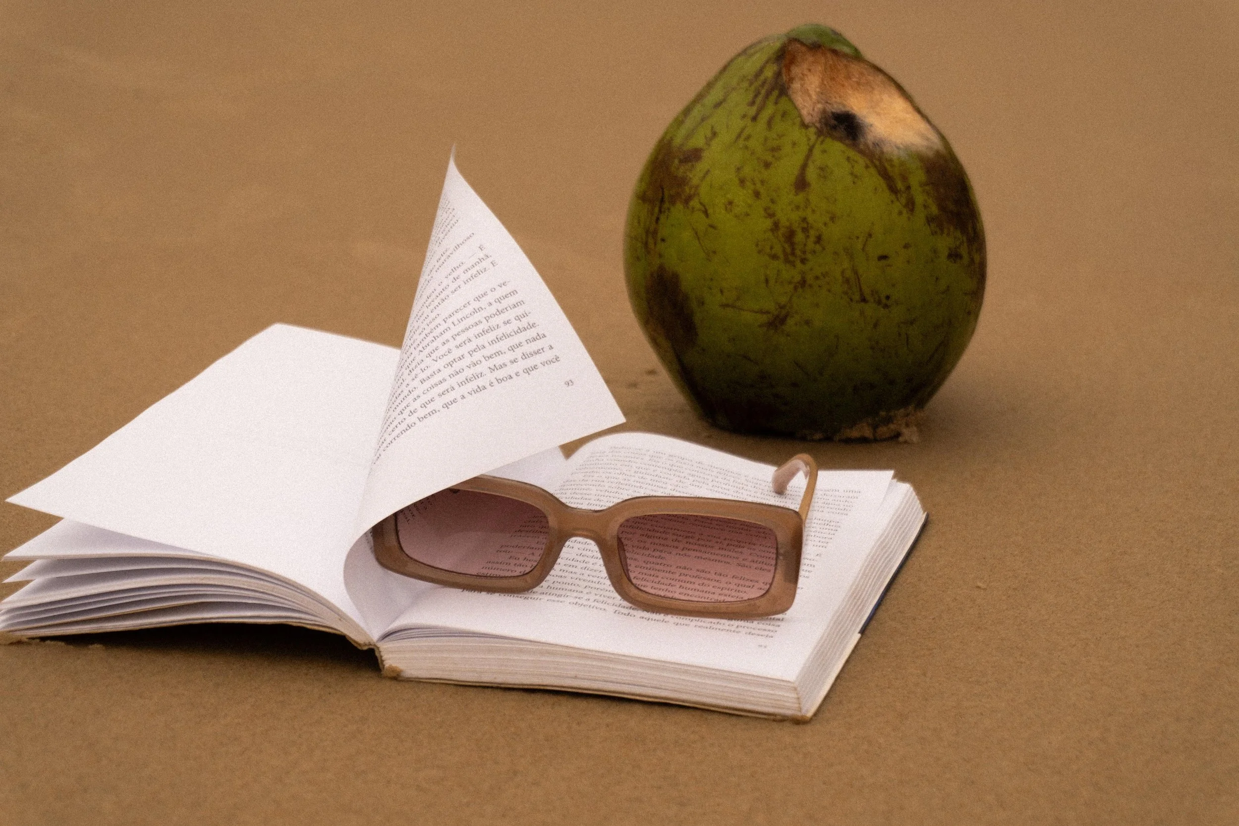 Open book with pink-tinted sunglasses resting on its pages, a green coconut with a brown patch in the background, all on a brown surface.