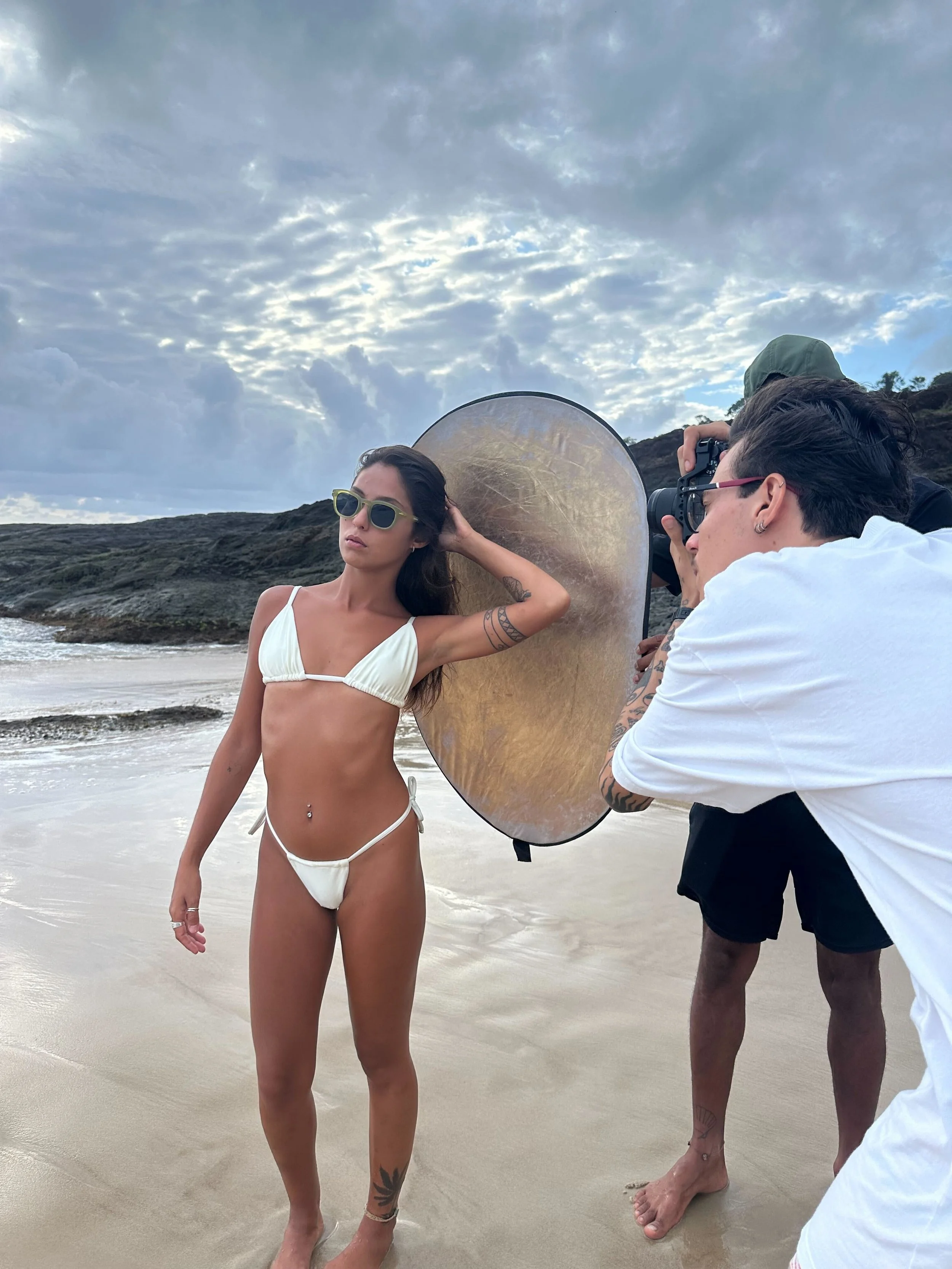 A model posing in a white bikini on the beach during a photo shoot, with a photographer taking her picture and an assistant using a light reflector, under cloudy skies.