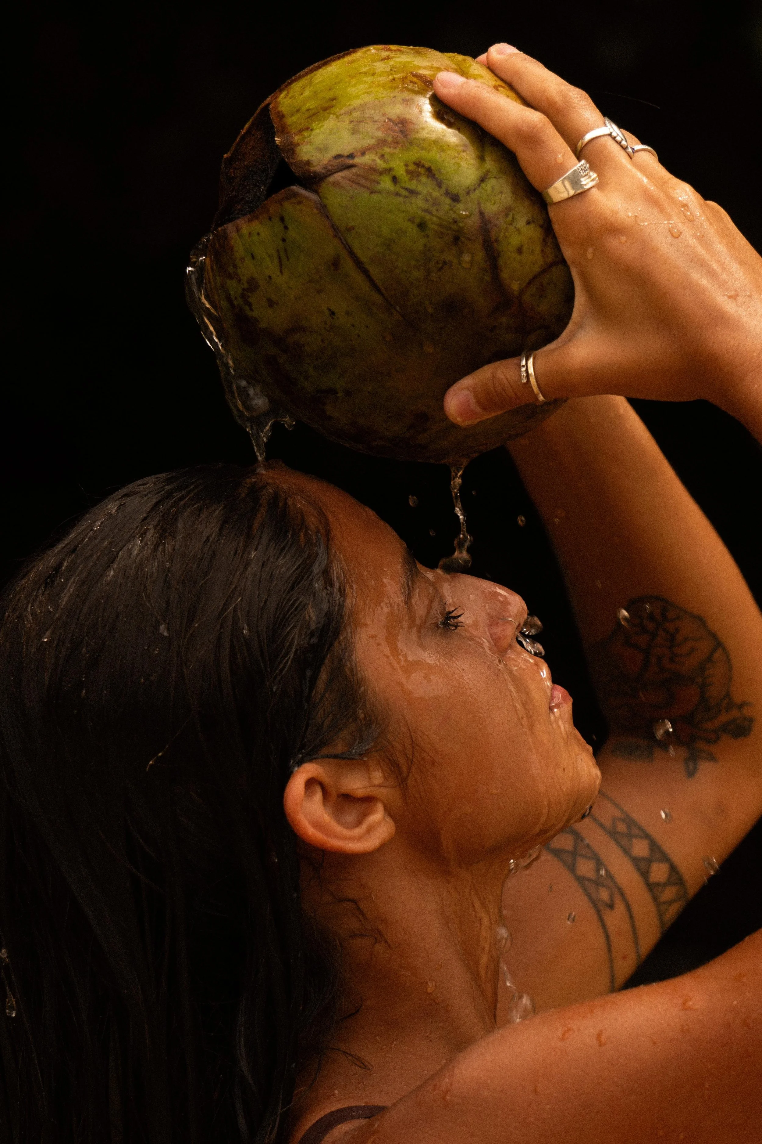 A woman with dark hair is pouring water from a green coconut onto her face and head.