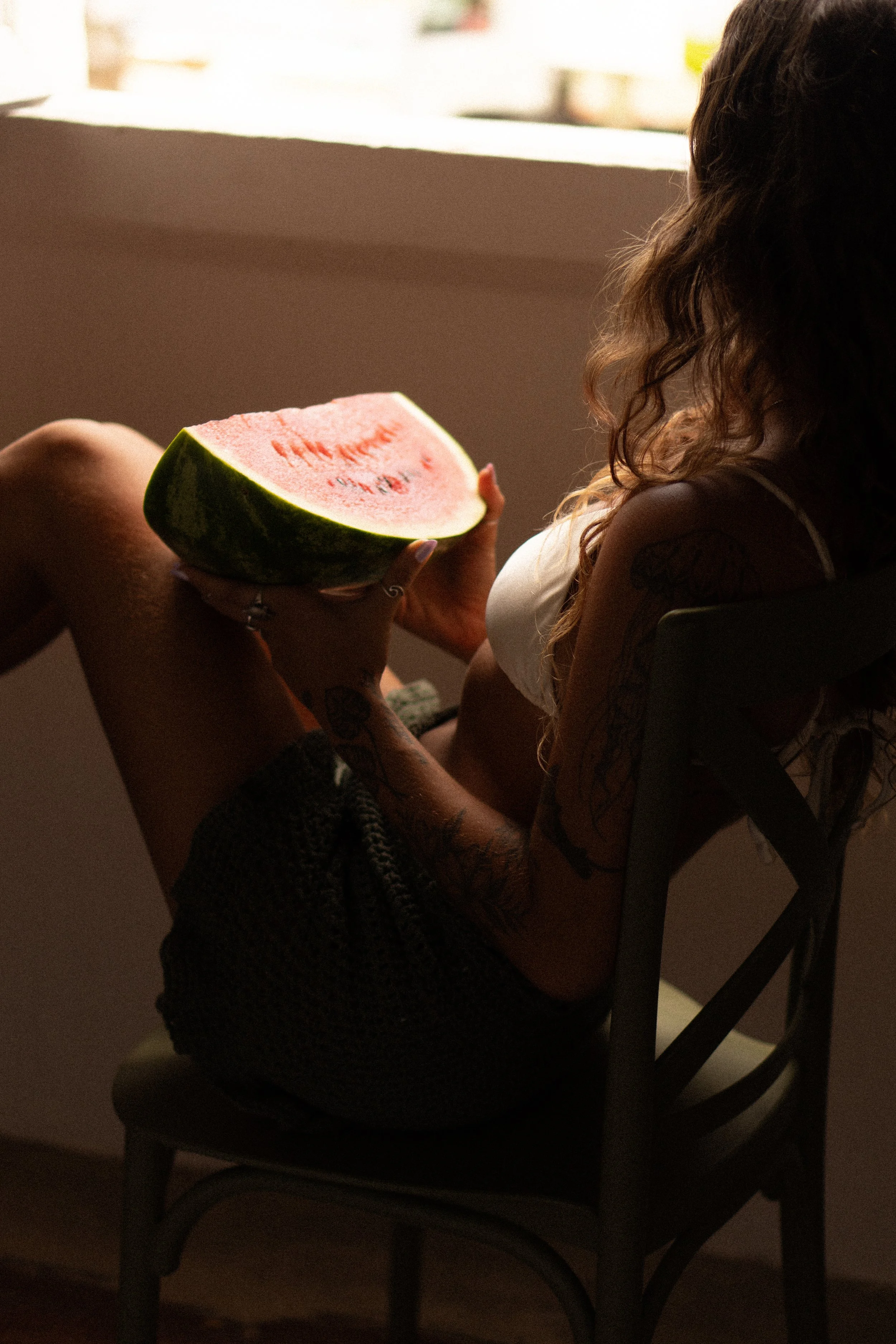 A woman with tattoos sitting in a chair, holding a slice of watermelon, in a warmly lit room near a window.