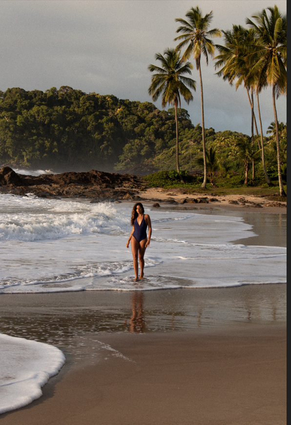 A woman in a navy swimsuit walking in shallow water on a sandy beach with tropical palm trees and lush green hills in the background.