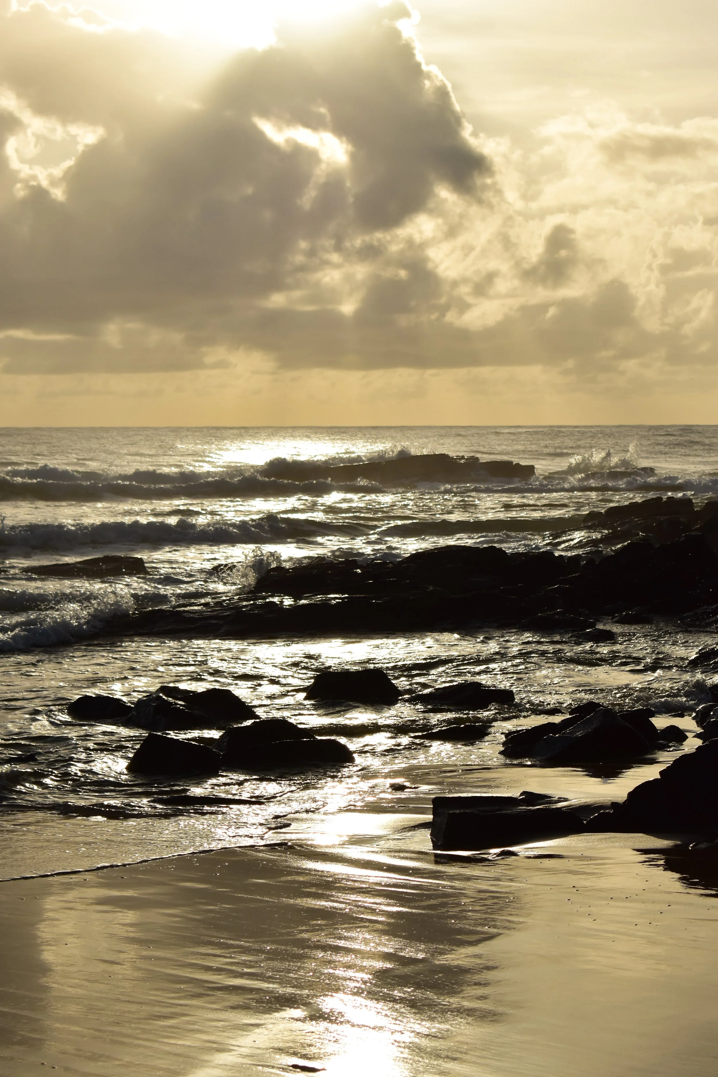 Sunset over a rocky shoreline with waves crashing and reflecting light on wet sand.