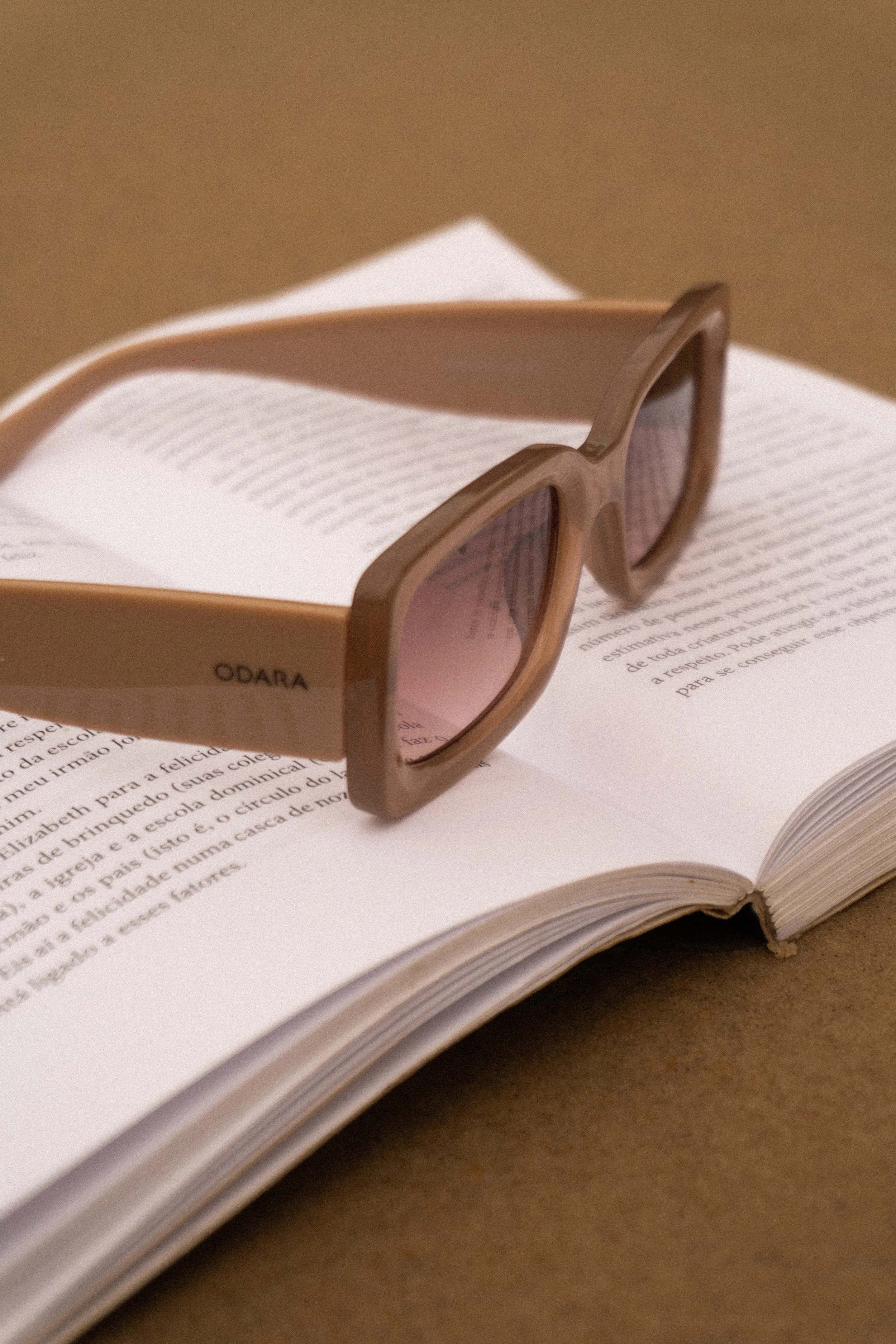 A pair of brown sunglasses resting on an open book with small print, on a brown surface.