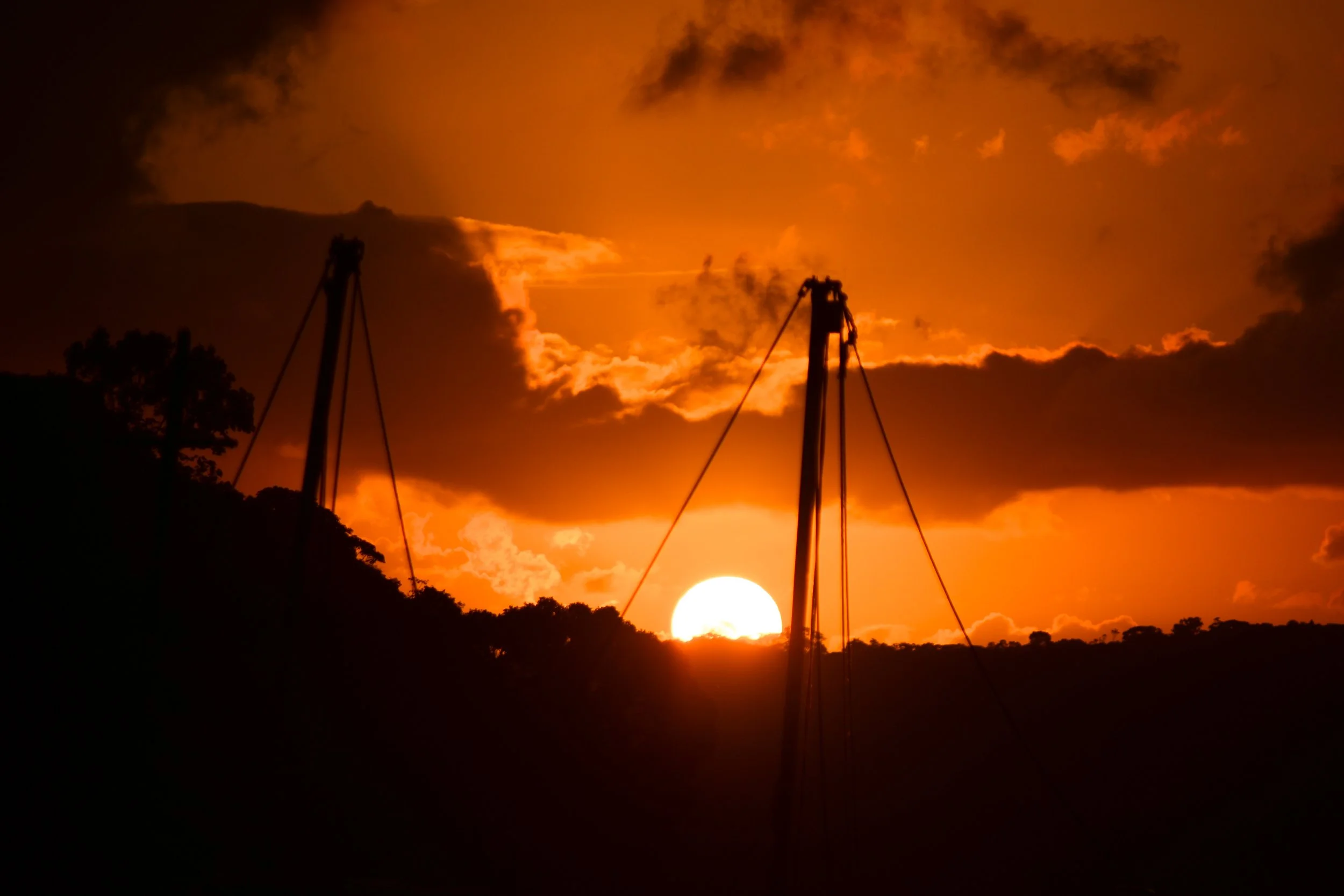 Sunset with the sun near the horizon, framed by dark silhouettes of two sailboat masts and a hilly landscape, with clouds in an orange sky.