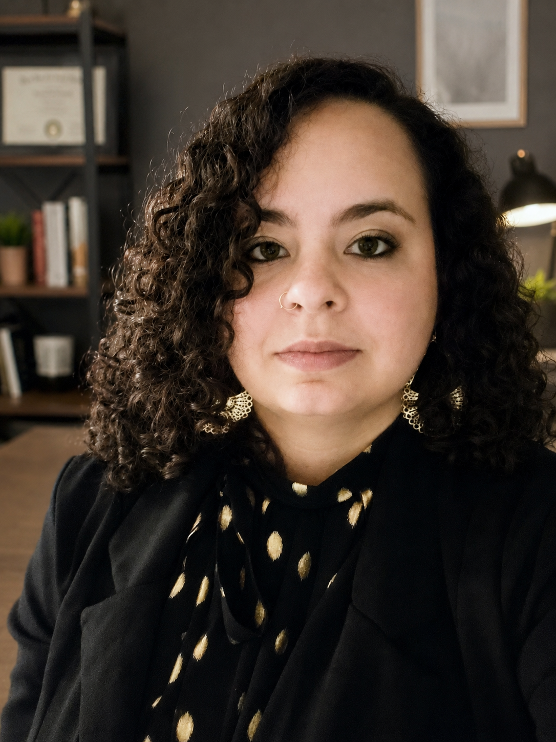 A woman with curly dark hair wearing a black top with gold polka dots, gold earrings, and a nose ring, in an indoor setting with a desk, bookshelf, and wall decorations in the background.