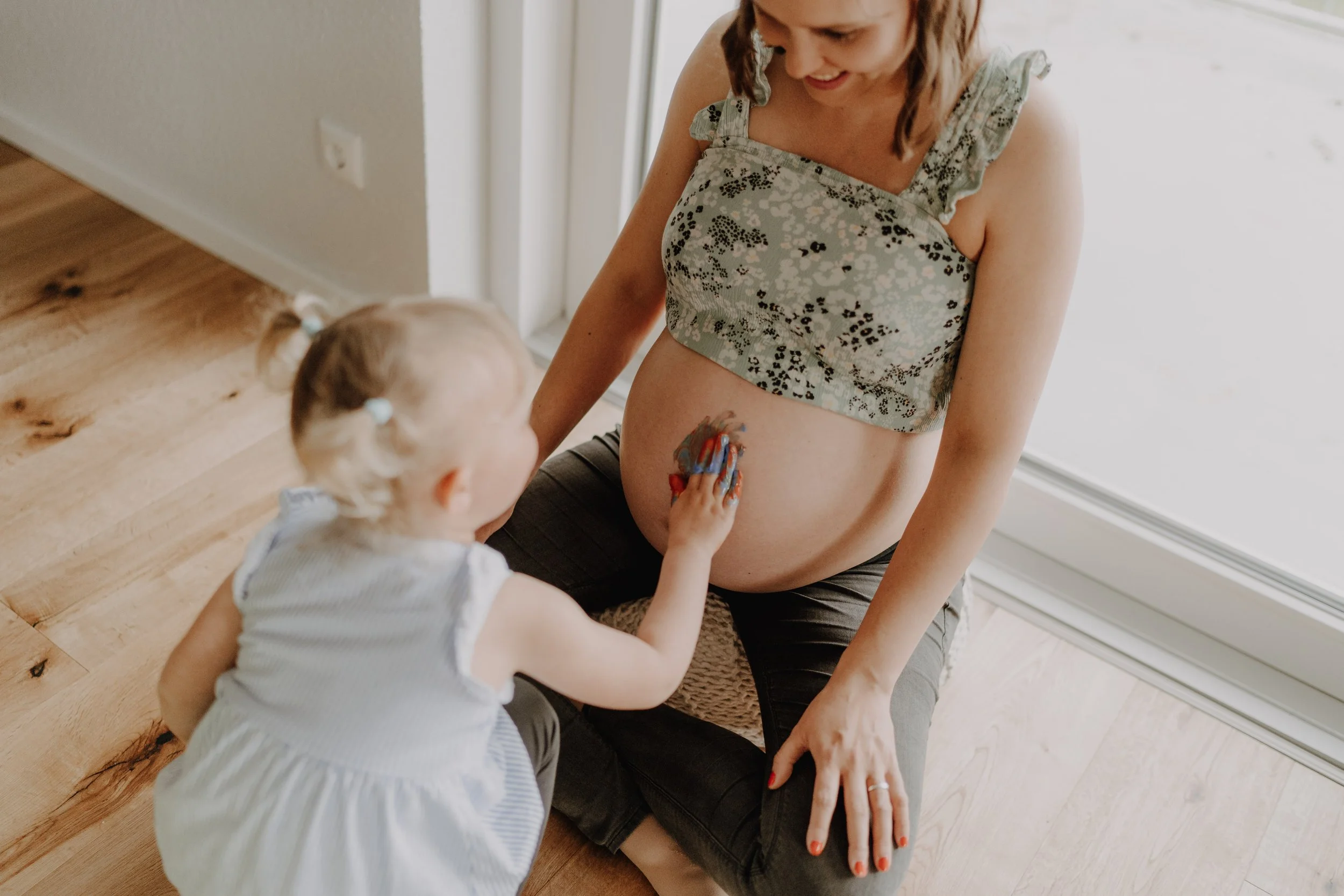 Eine schwangere Frau mit ihrer Tochter beim Babybauchshooting in Lemgo. Das Kind bemalt den Bauch der Mutter mit Fingerfarben. Fotostudio Claudia Hilgenböker Lemgo.