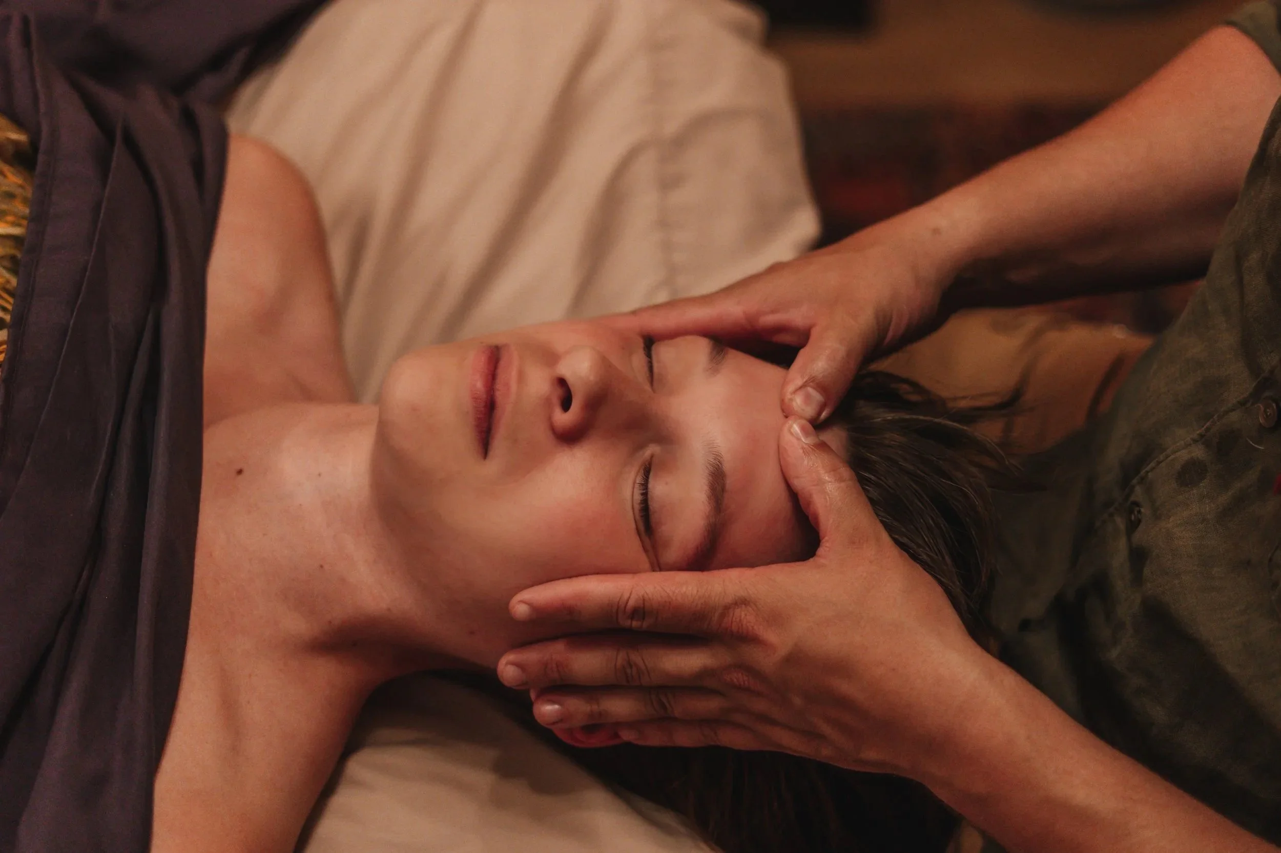 woman with eyes closed getting a massage laying down on a tan sheet. massage therapist has two hands on her head, applying pressure with his thumbs on the center of her forehead.