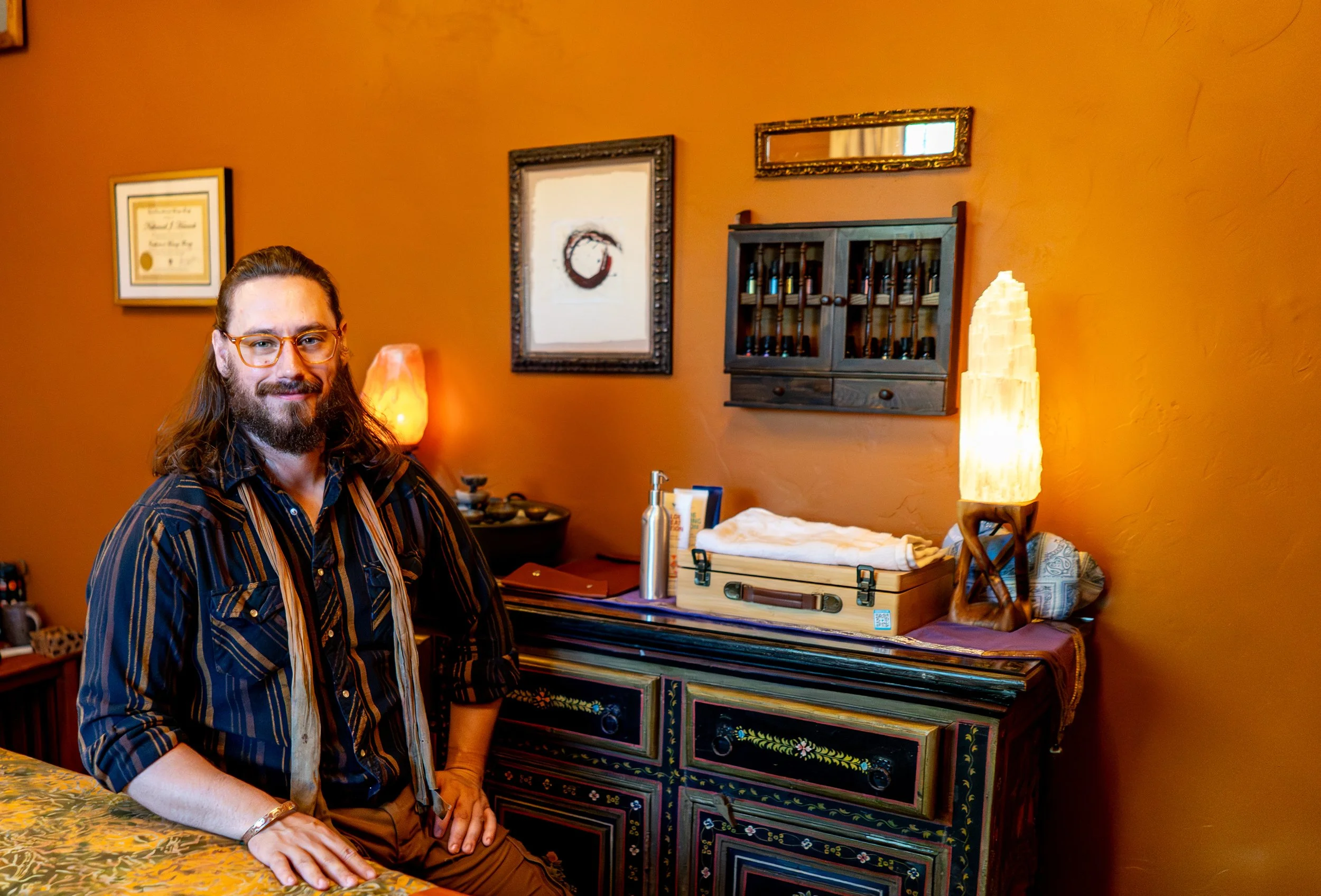 man with orange framed glasses that has a full beard and medium length brown hair. Wearing a striped black and tan button down shirt, sitting at a table. orange wall behind him.