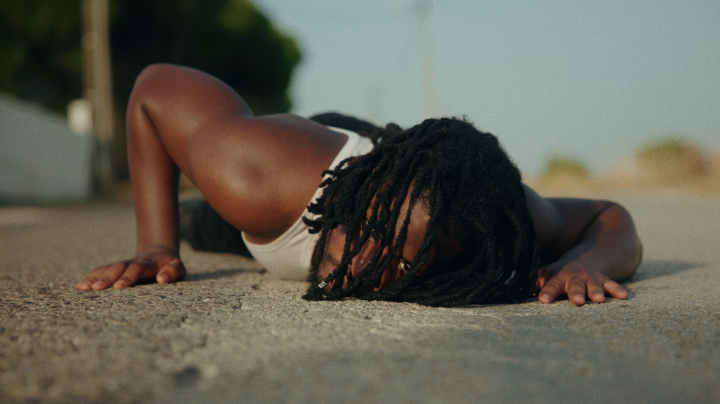 A person with dreadlocks performs a push-up on a sandy surface outdoors, wearing a sleeveless top.