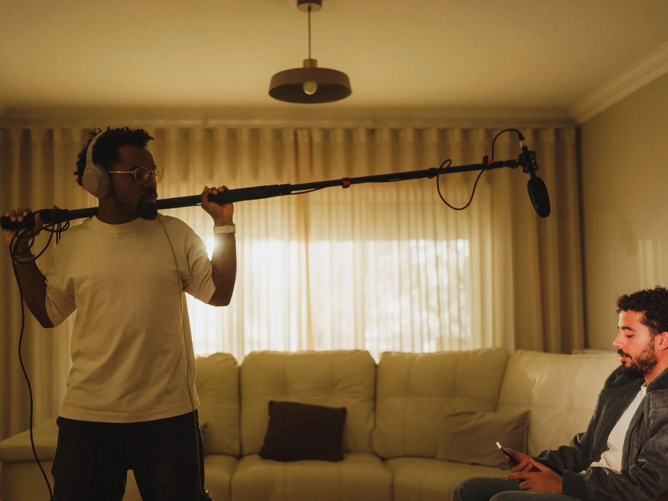 A man wearing headphones and glasses holds a boom microphone over his shoulder while recording. Another man sitting on a couch in the living room uses a smartphone.