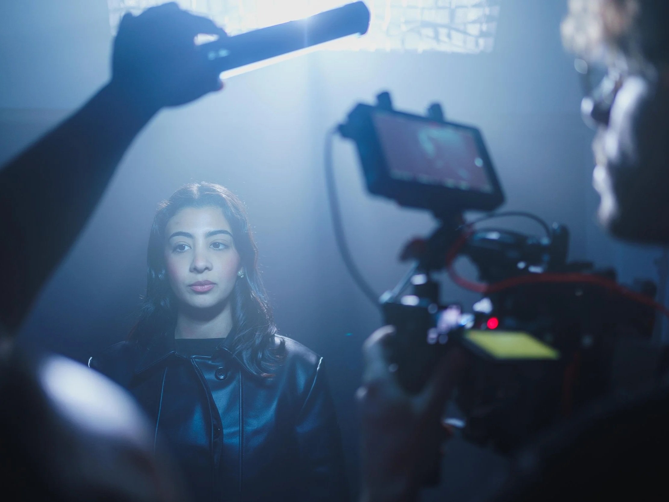 A woman with dark hair sits in front of a camera on a film set, with a crew member adjusting the equipment in the foreground. The scene is illuminated with soft, dramatic lighting.